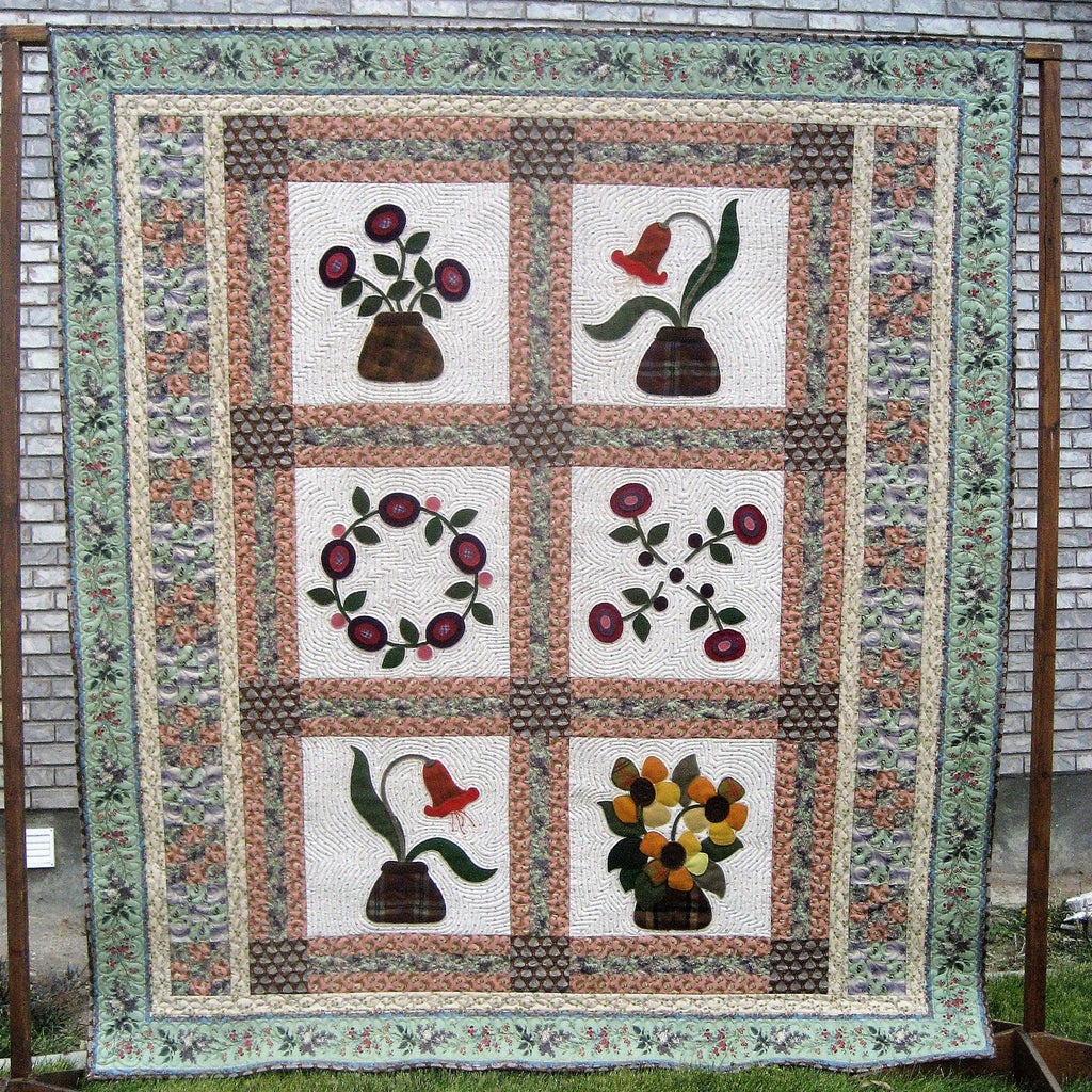 A vintage-style floral quilt hanging outdoors on a wooden frame against a gray brick wall and green grass. The quilt features six square panels with different floral and botanical designs, surrounded by a patterned border.