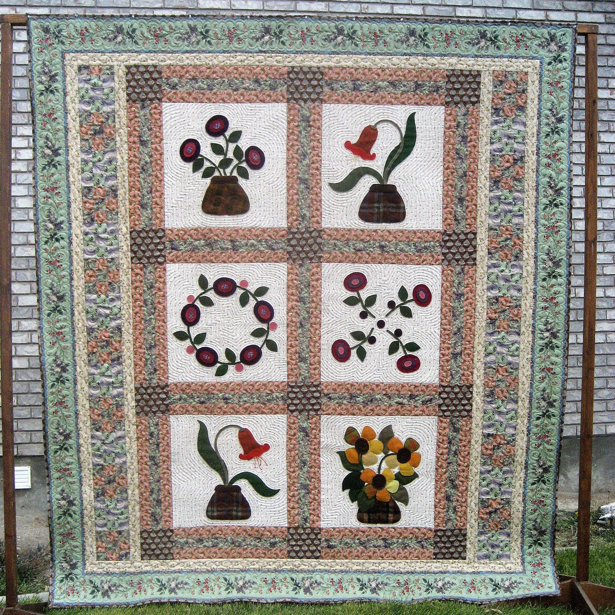 A vintage-style floral quilt hanging outdoors on a wooden frame against a gray brick wall and green grass. The quilt features six square panels with different floral and botanical designs, surrounded by a patterned border.