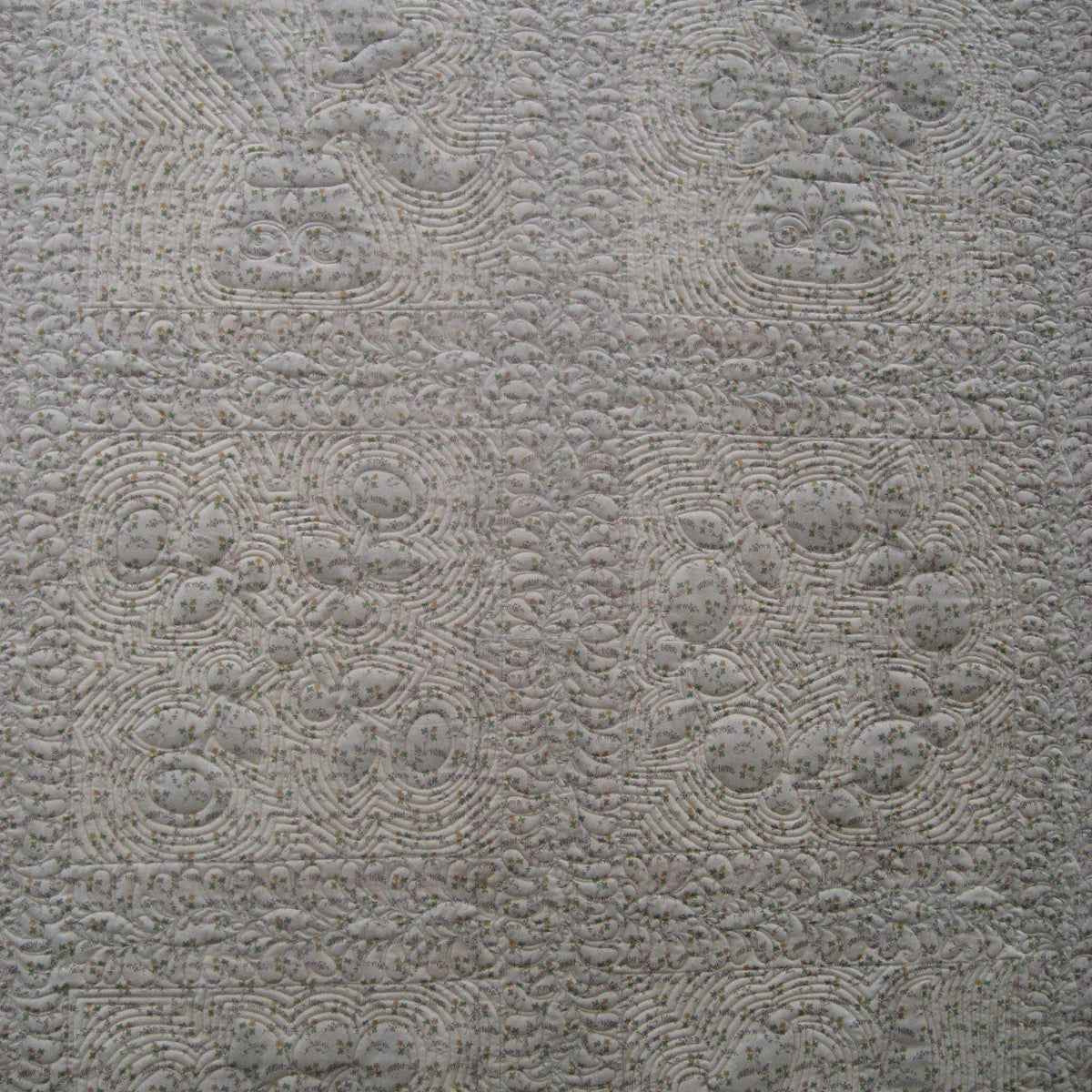 A close-up of a light gray quilted fabric with a repeating floral and scroll pattern.