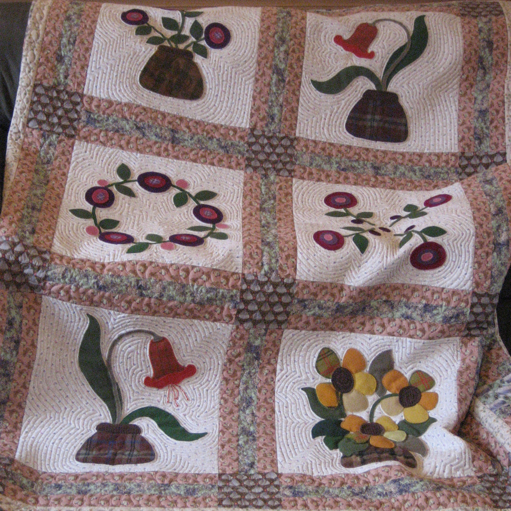 A handmade quilted patchwork blanket featuring six floral and botanical panels with appliques of flowers, leaves, and berries, bordered by a floral patterned fabric.