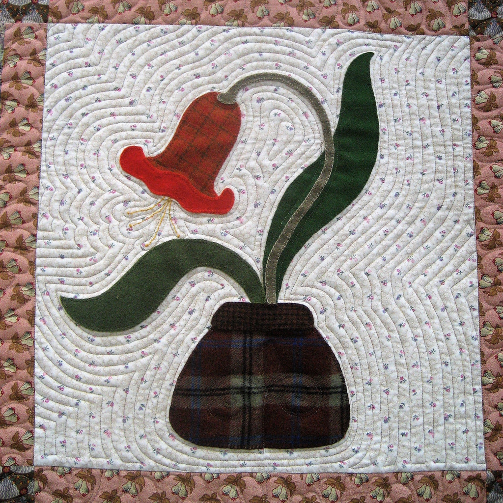 A close-up of a quilted textile featuring a stylized tulip applique in a dark tartan pot, with green leaves and a red flower, set against a white wavy background and surrounded by a pink floral border.