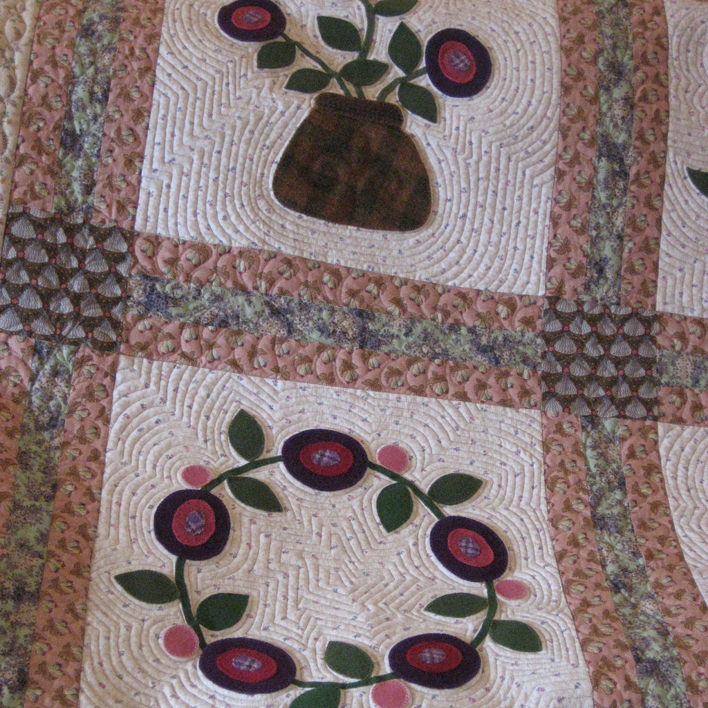 A close-up of a quilted textile featuring cream square panels with floral and botanical applique designs, including a vase with flowers at the top and a wreath of red and purple flowers with green leaves at the bottom, bordered by pink and brown patterned fabric.