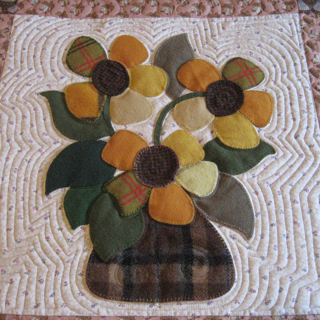 A handmade quilted fabric applique featuring two yellow and orange sunflowers with green leaves in a brown plaid vase, set against a white quilted background with a pink floral border.