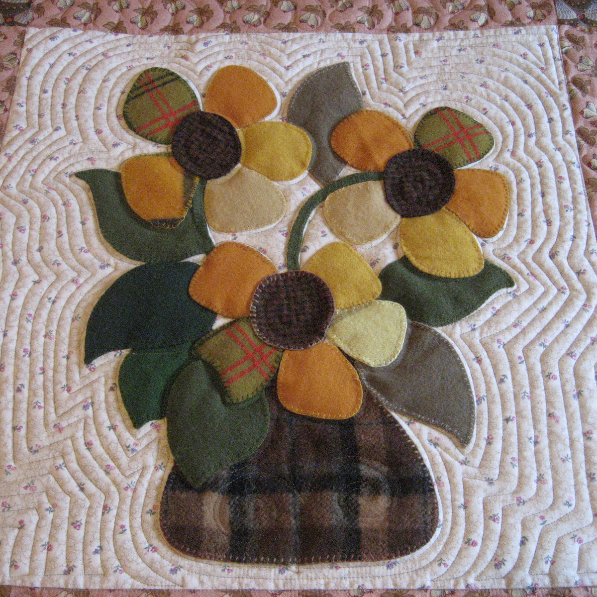 A handmade quilted fabric applique featuring two yellow and orange sunflowers with green leaves in a brown plaid vase, set against a white quilted background with a pink floral border.