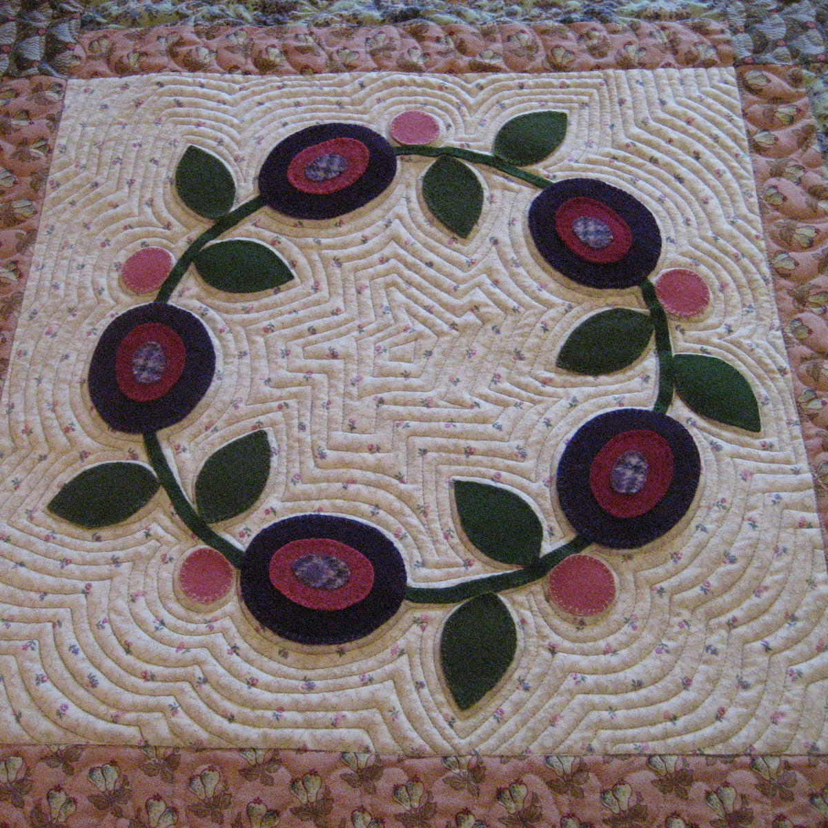 A close-up of a quilted fabric panel featuring a circular floral wreath design with dark purple and red flowers, green leaves, and pink accents on a cream background.