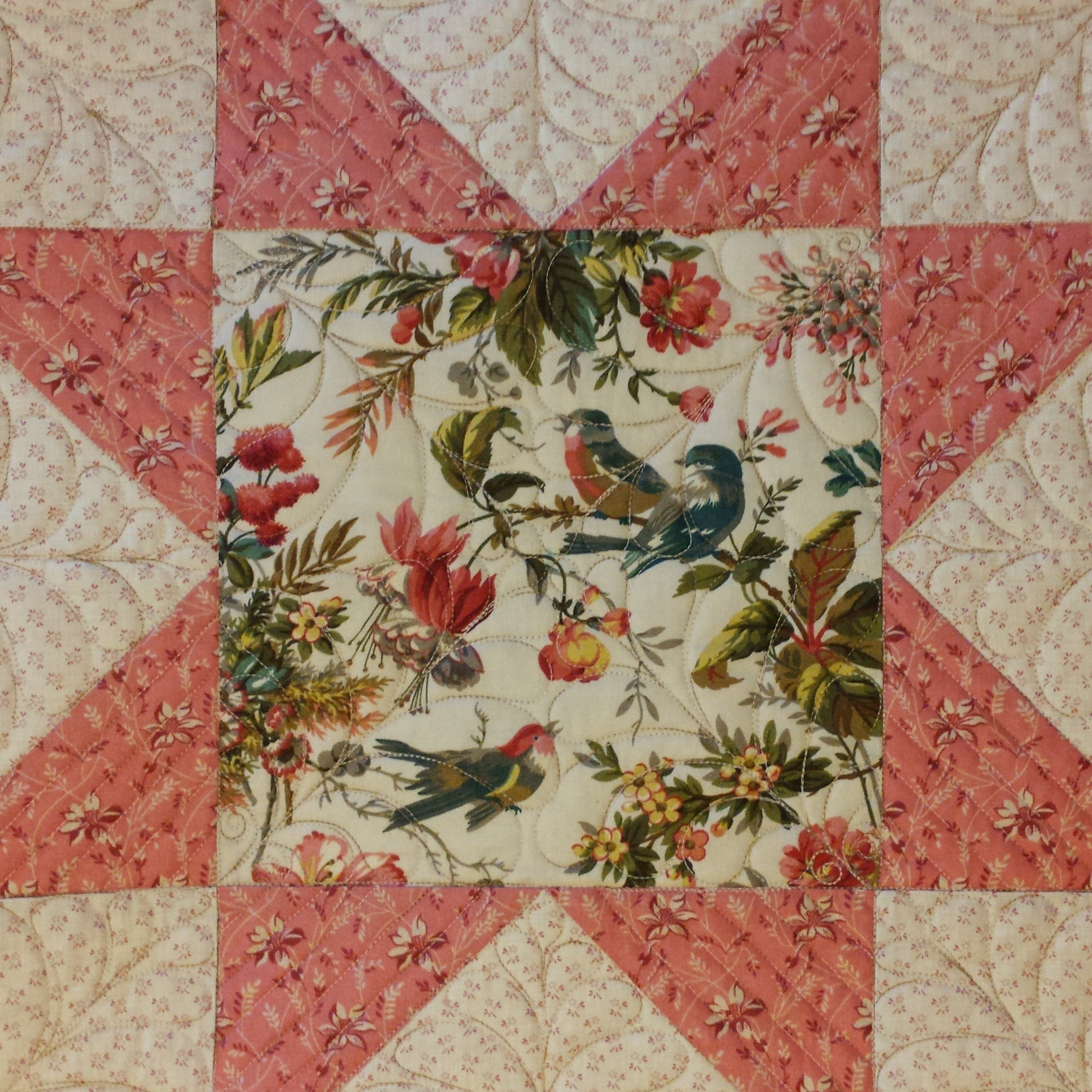 A close-up of a vintage-style quilted textile featuring a central floral and bird print surrounded by coral starburst sections and cream patterned borders with burgundy flowers.