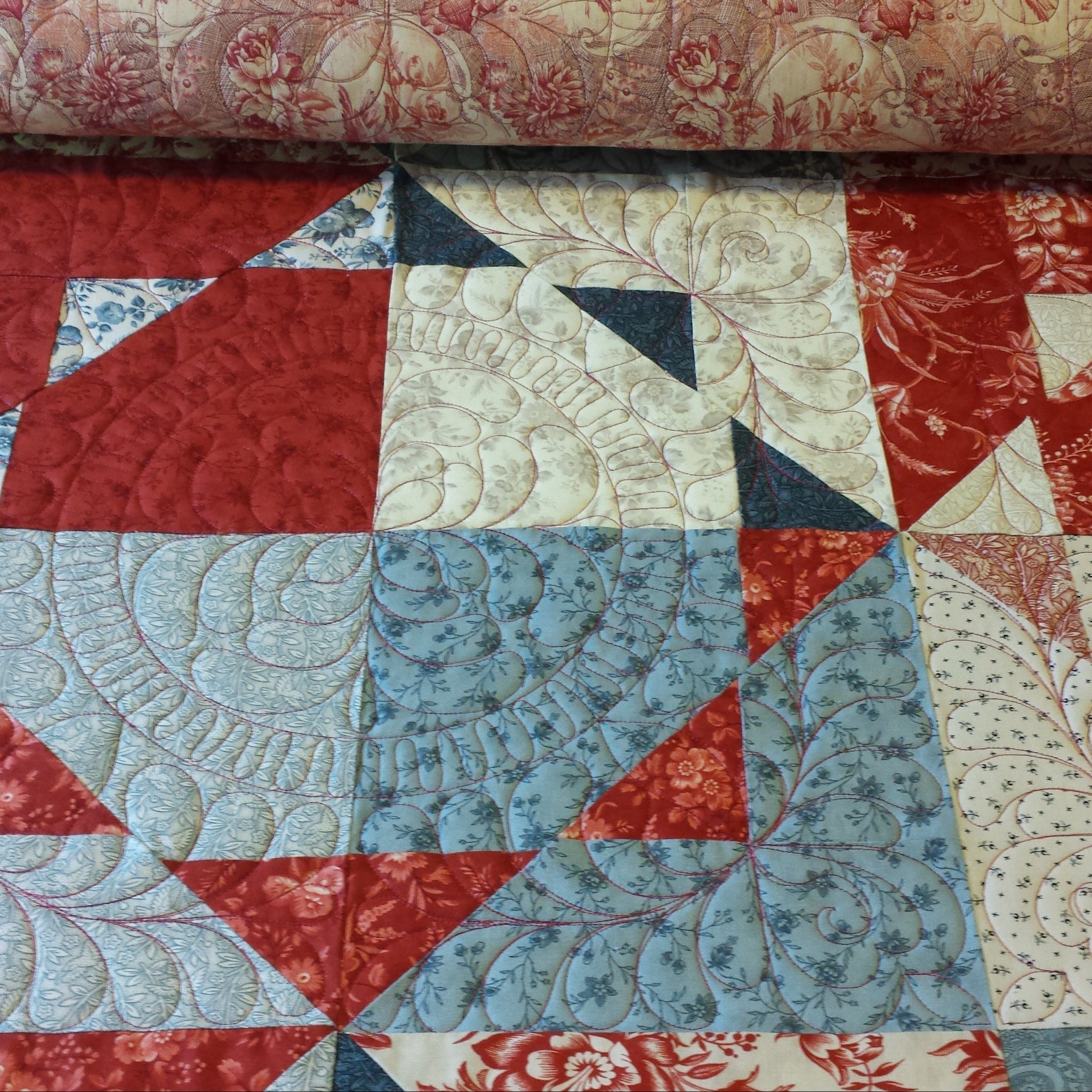 A close-up of a quilted fabric with a geometric patchwork design in red, light blue, and cream, with a matching patterned fabric roll visible at the top.