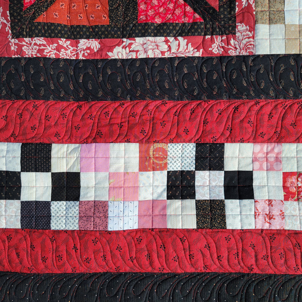 A close up of a quilt with a variety of floral prints in black and red with a checkerboard strip in the middle. 