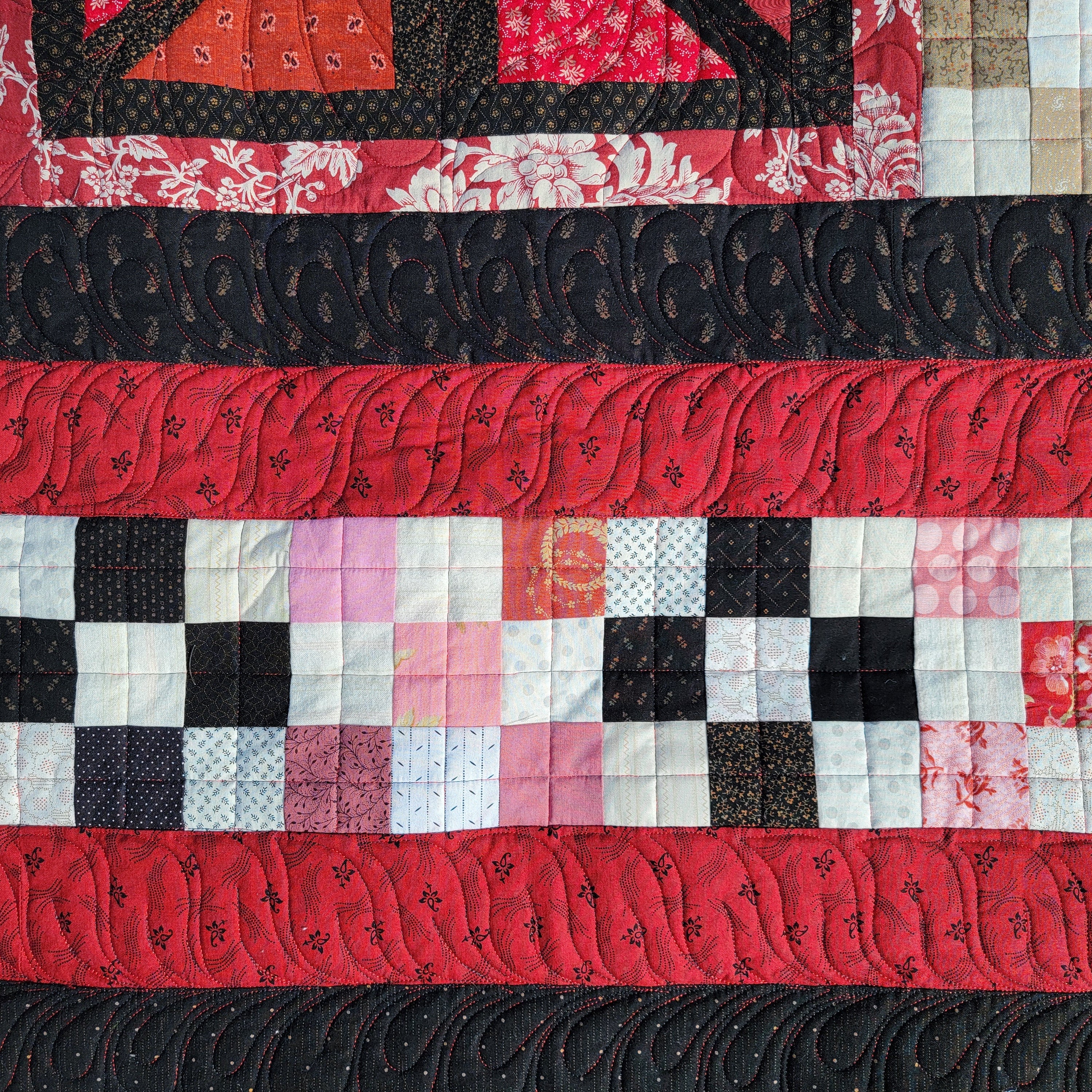 A close up of a quilt with a variety of floral prints in black and red with a checkerboard strip in the middle. 