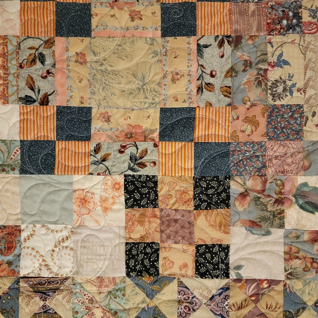 A vintage patchwork quilt featuring a grid of square and diamond blocks in various floral, striped, and solid fabrics. The color palette is a mix of dusty pink, navy blue, beige, and muted orange, with some blocks showing embossed floral patterns.