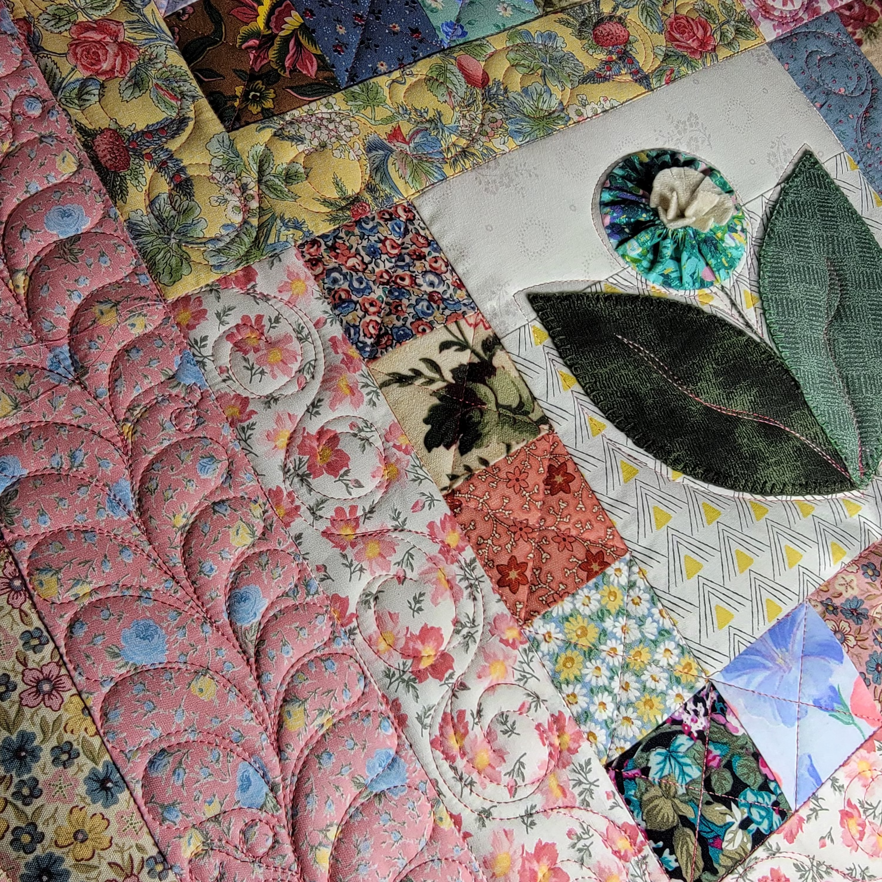 A close-up of a colorful patchwork quilt with floral and geometric fabrics, featuring a prominent pink scalloped section and a white square patch with green leaf embroidery.