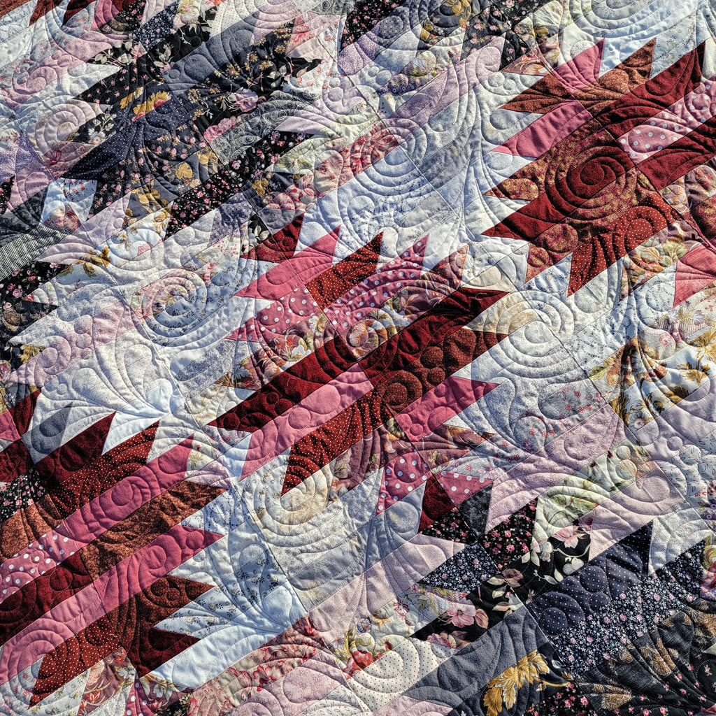 A close-up of a quilted patchwork textile with repeating geometric and floral fabric blocks in pink, burgundy, black, white, and light blue, arranged in diagonal rows with swirling quilting patterns.