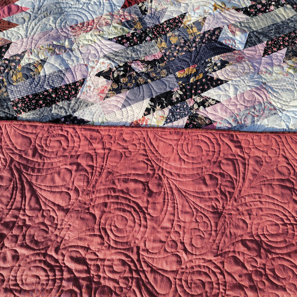 Close-up of a quilted textile with a dusty rose pink lower section featuring swirling floral quilting, and a multicolor patterned upper section with star and geometric patchwork designs.