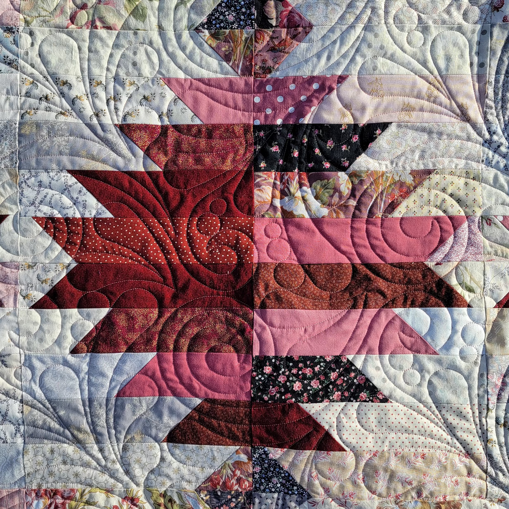 A close-up of a quilted fabric panel featuring a geometric starburst pattern in shades of red, pink, black, and white, with detailed swirling quilting stitched across the textile.