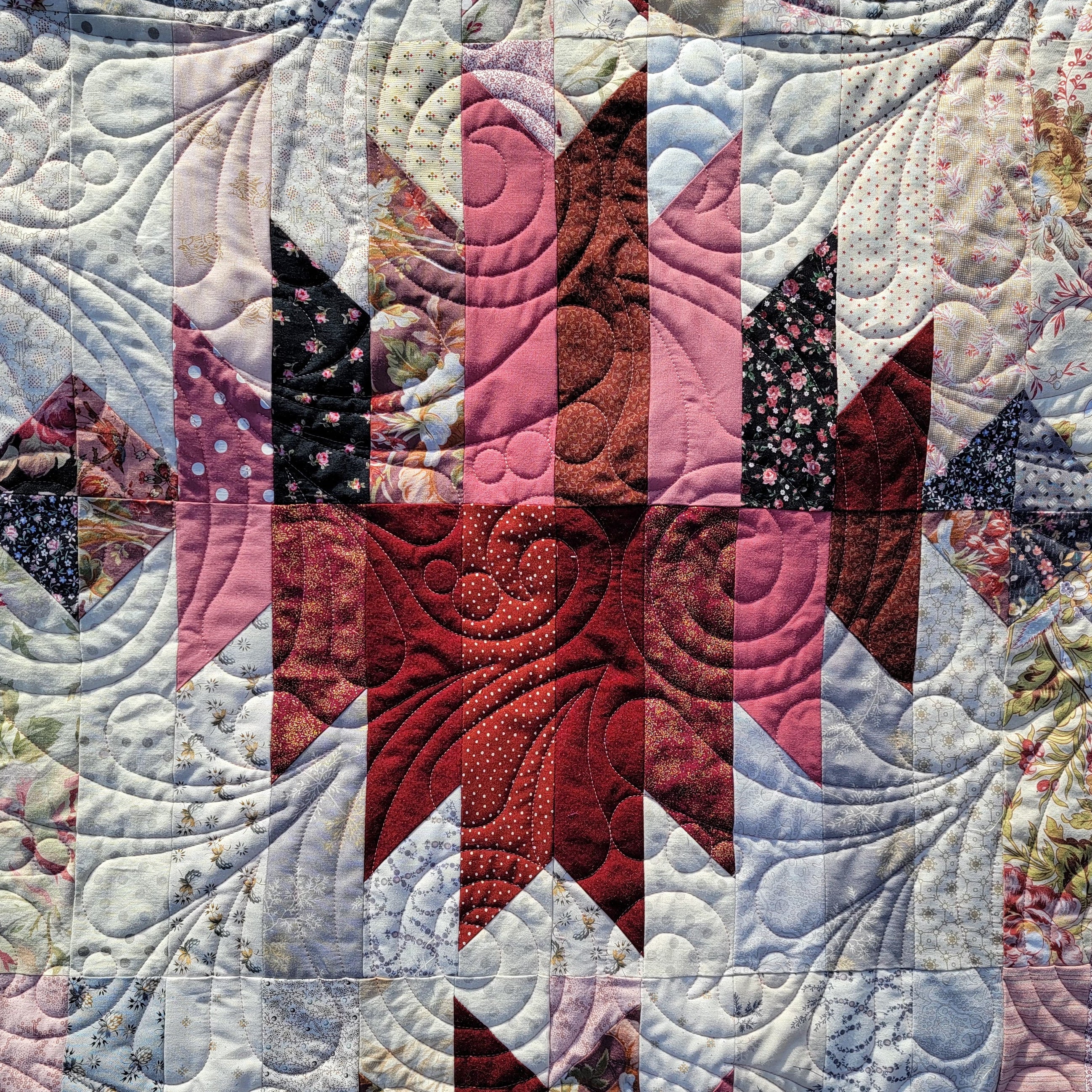 A close-up of a quilted fabric featuring a geometric starburst pattern in shades of red, pink, and dark navy, surrounded by light blue and white floral and paisley fabrics with intricate swirling quilting.