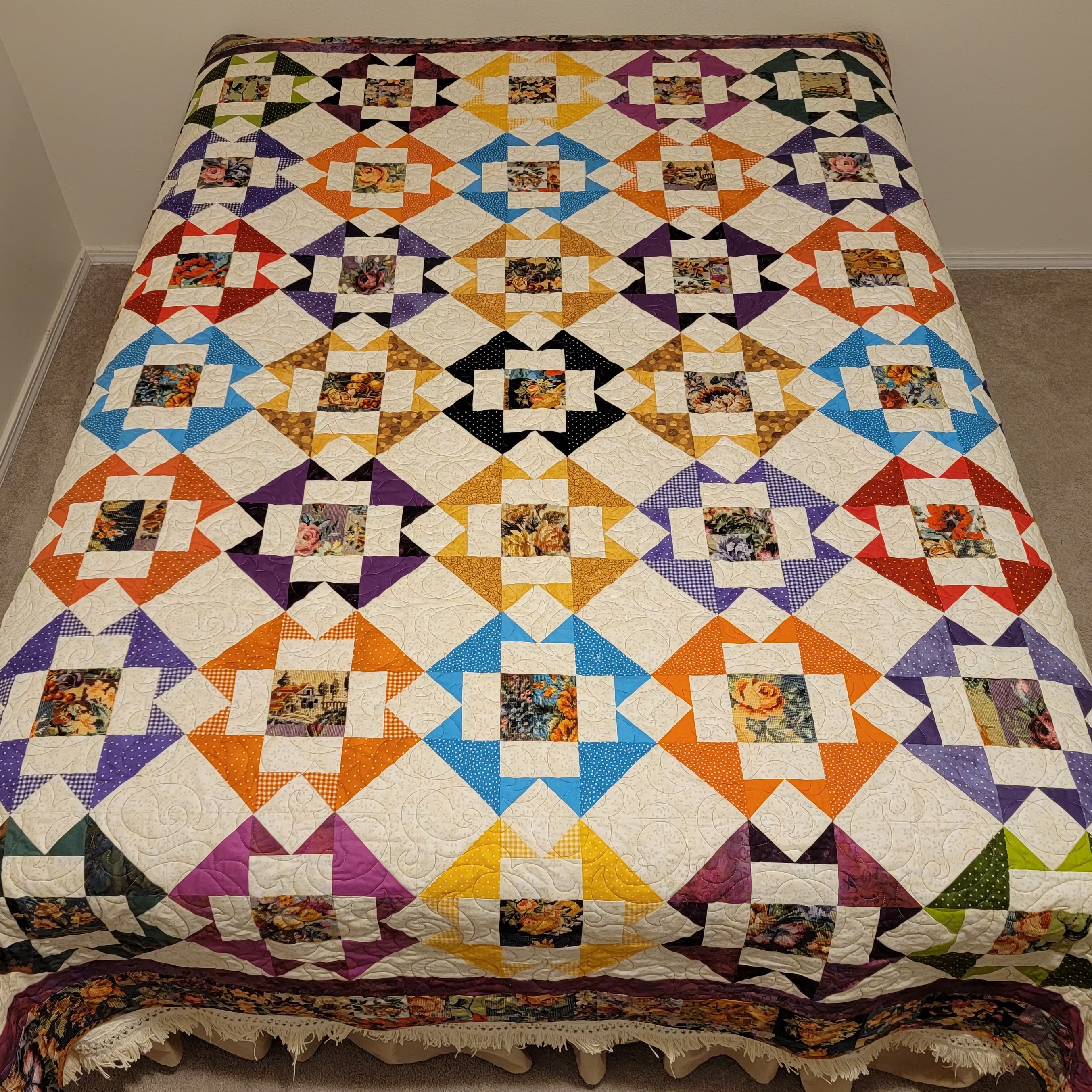 A colorful patchwork quilt with a geometric diamond pattern and floral center squares, spread out on a bed in a simple room with beige walls and carpet.