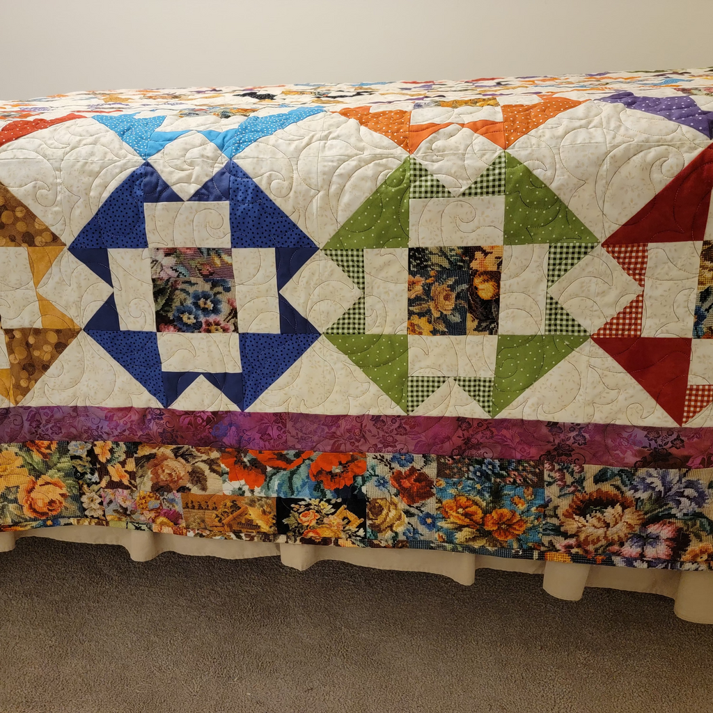 A colorful quilted bedspread or quilted cover with a geometric star pattern and floral border, draped over a bed in a bedroom setting.