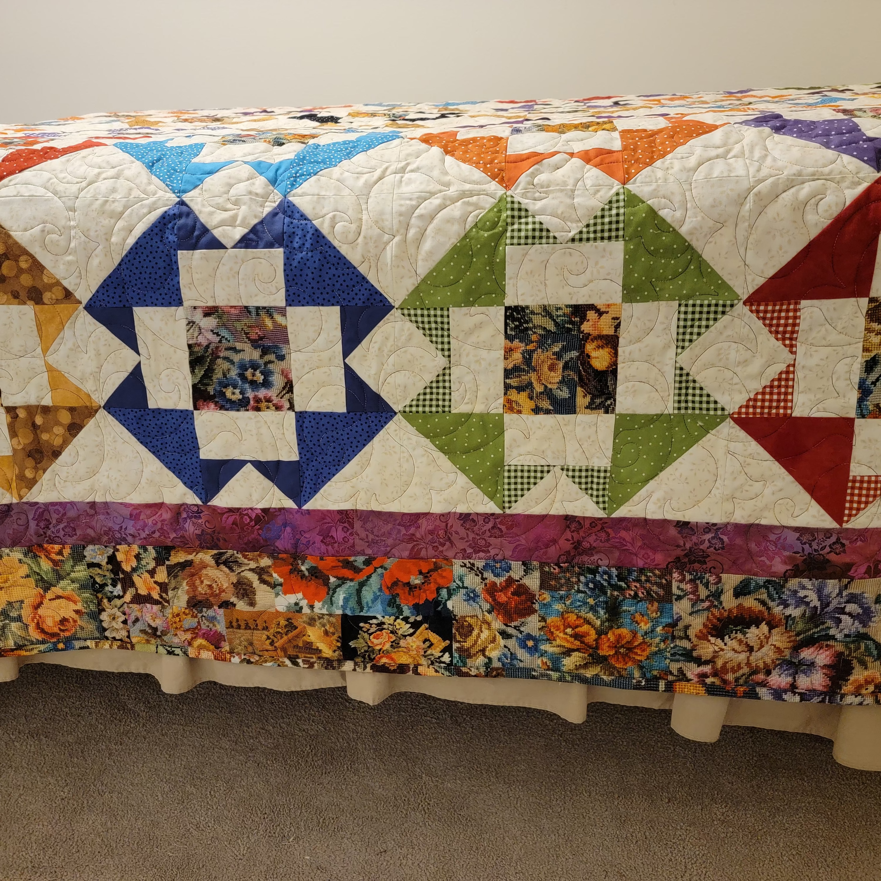 A colorful quilted bedspread or quilted cover with a geometric star pattern and floral border, draped over a bed in a bedroom setting.