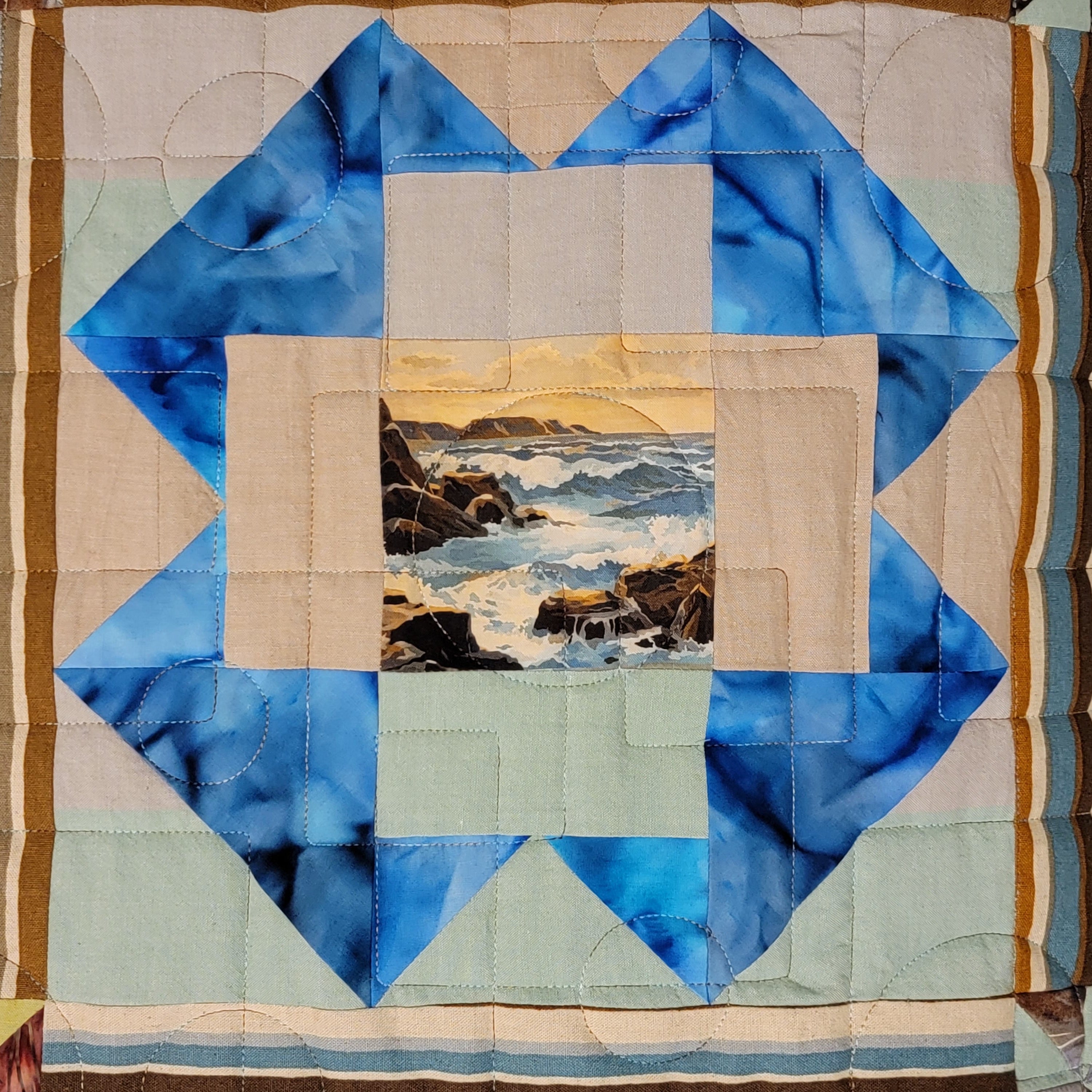 A square quilted textile featuring a geometric star-like border made of blue and beige fabric sections, with a central landscape patch showing ocean waves and rocky coastline. Striped border panels in beige, brown, and blue frame the quilt.