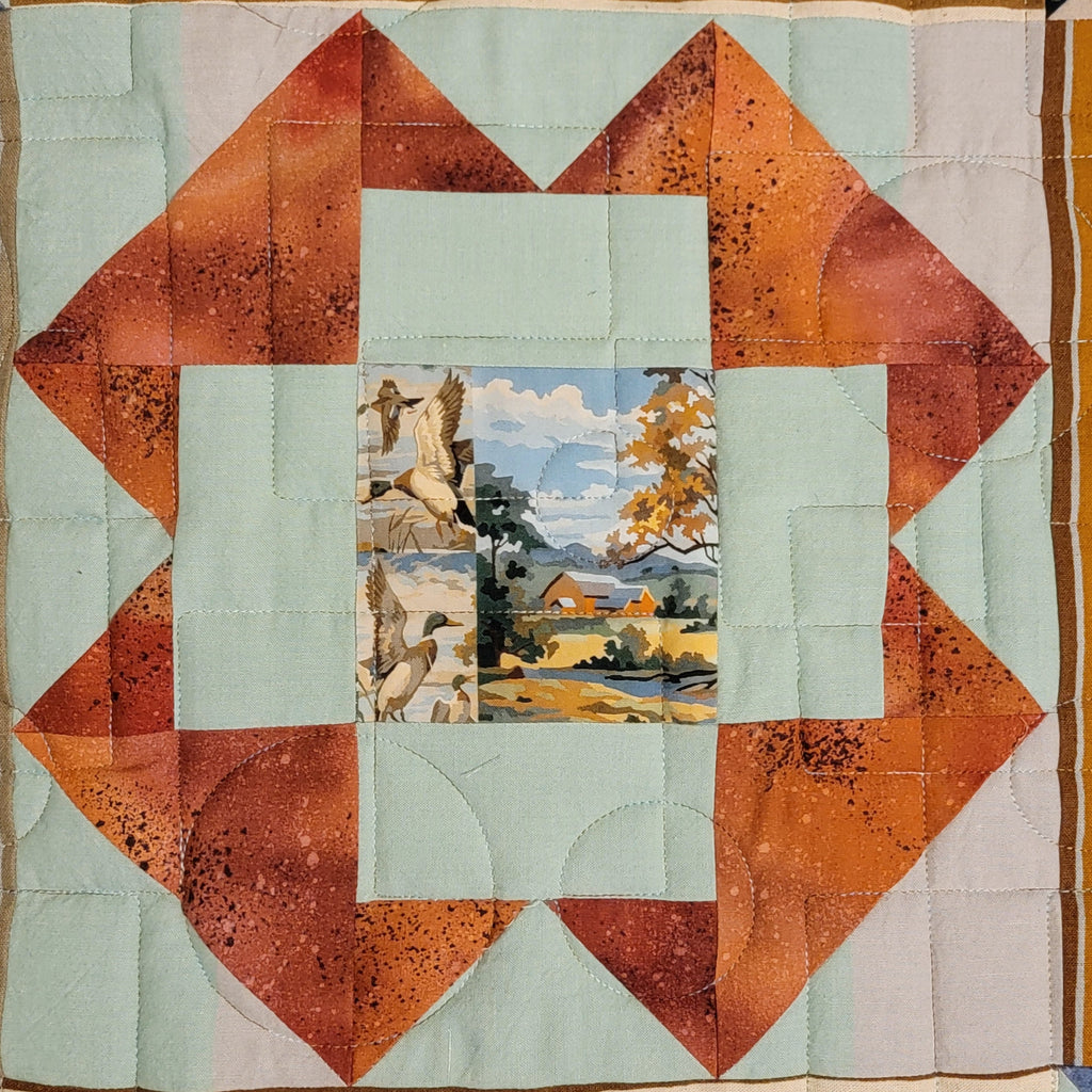 A quilted fabric piece featuring a geometric starburst pattern with orange and rust-brown triangles, light blue centers, and a small landscape patch in the middle.