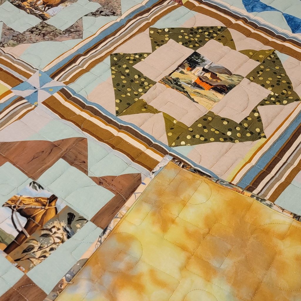 A close-up of a quilted patchwork textile with geometric blocks, striped borders, and small printed fabric inserts. The visible color story includes beige, brown, blue, olive green, and a large warm yellow-gold section in the lower right.