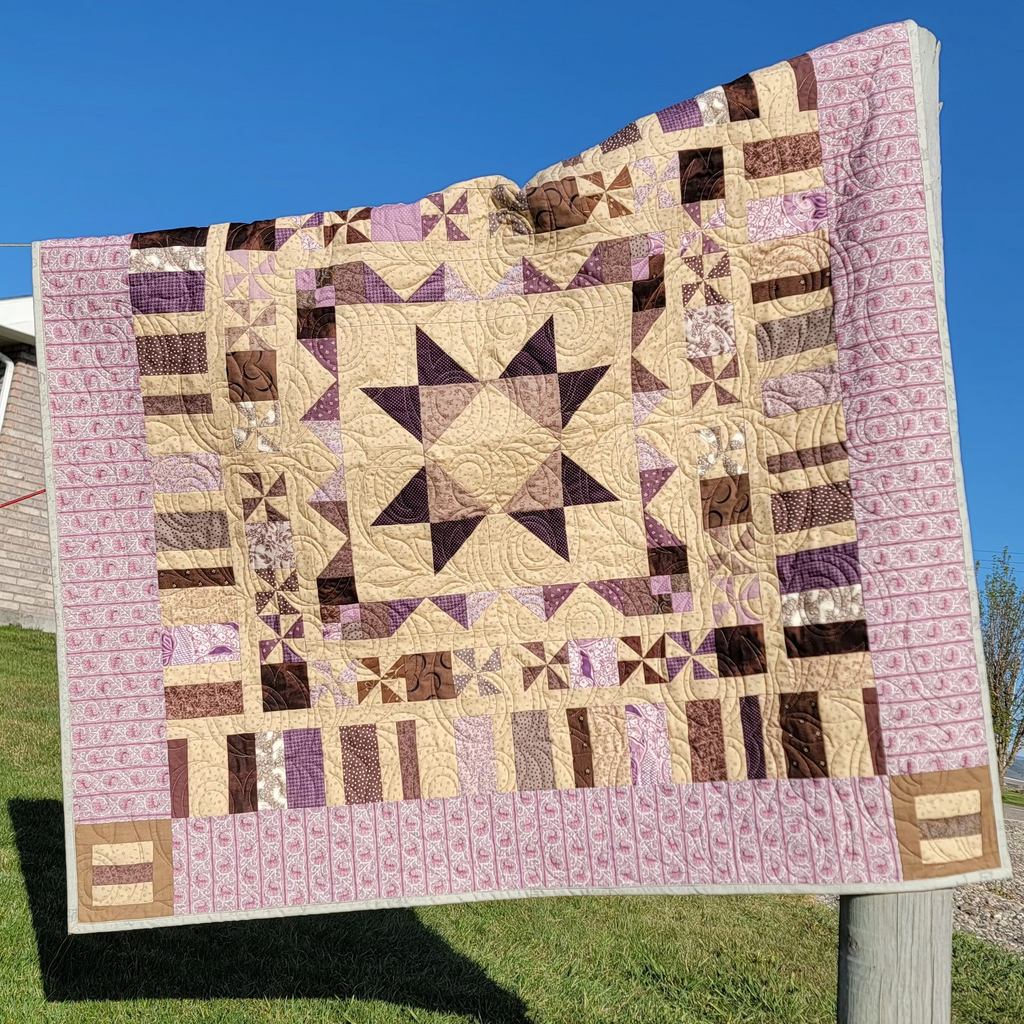 A handmade patchwork quilt with a geometric star pattern in shades of purple, brown, and beige, hanging outdoors on a wooden post against a blue sky and green grass background.