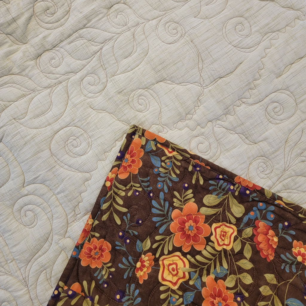 A close-up of a quilted cream-colored blanket with swirling stitched patterns, showing the reverse side in the bottom corner. The reverse side features a dark brown background with a vibrant floral print in orange, yellow, and green.