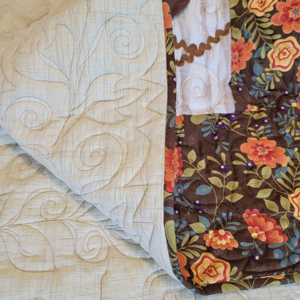 A close-up of a quilt, showing two distinct sections: one side in light beige with swirling stitched patterns, and the other side in a brown floral print with orange, red, and green flowers.