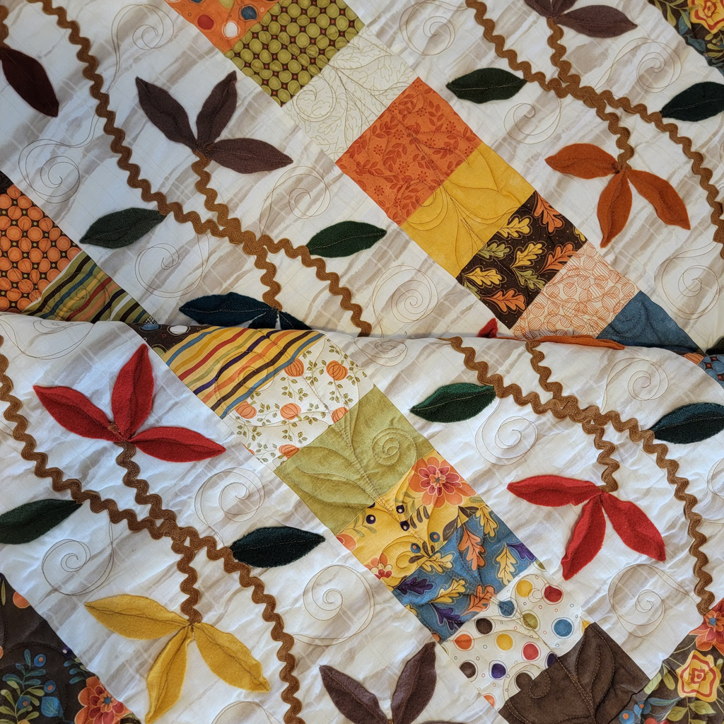 A close-up of a patchwork quilt featuring autumn-themed leaf appliqués in red, yellow, brown, and green, set against a cream background with wavy quilting stitches and colorful fabric squares.