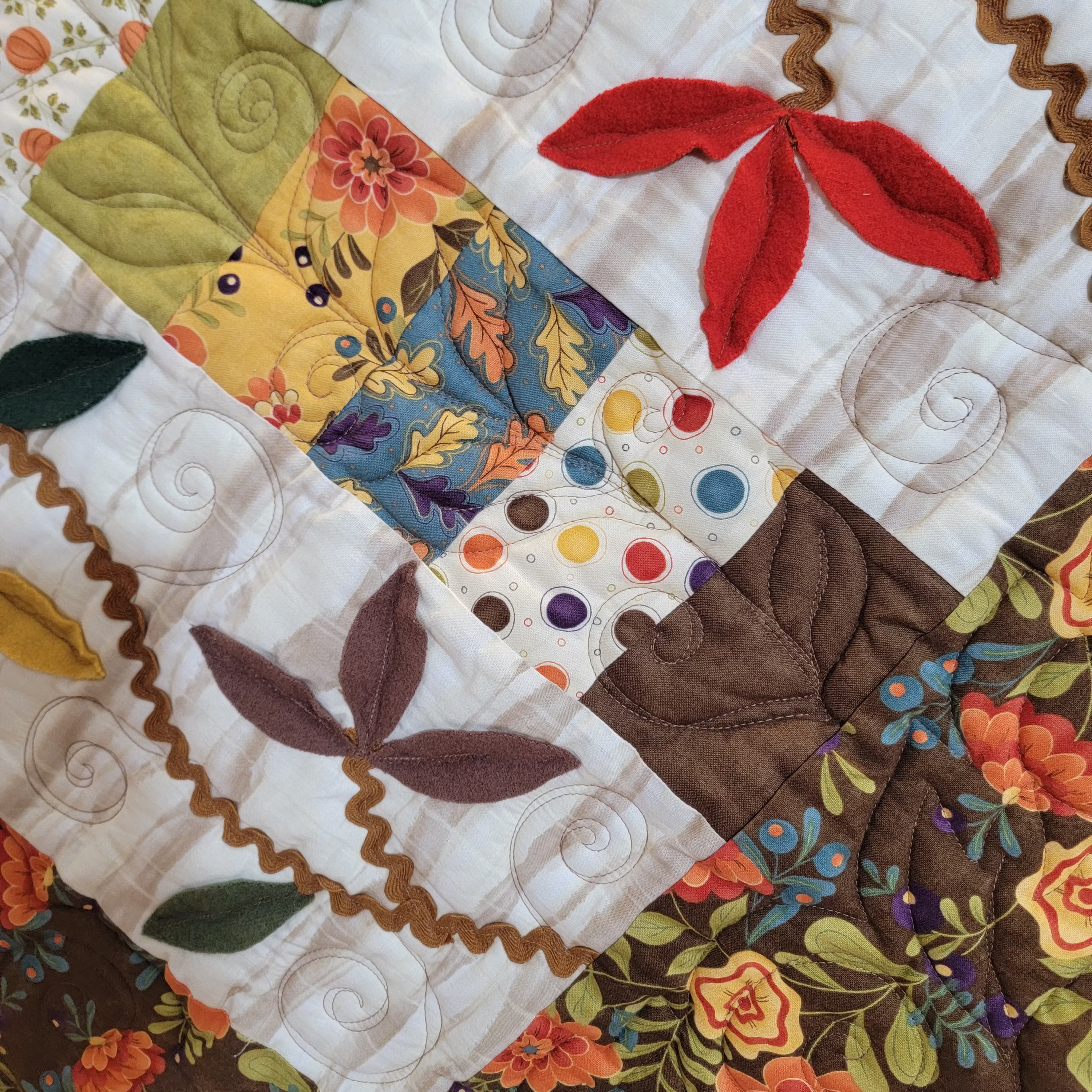 A close-up of a patchwork quilt featuring various fabric patterns, including floral prints, polka dots, and embroidered leaf motifs in autumn colors like orange, red, green, yellow, and brown.