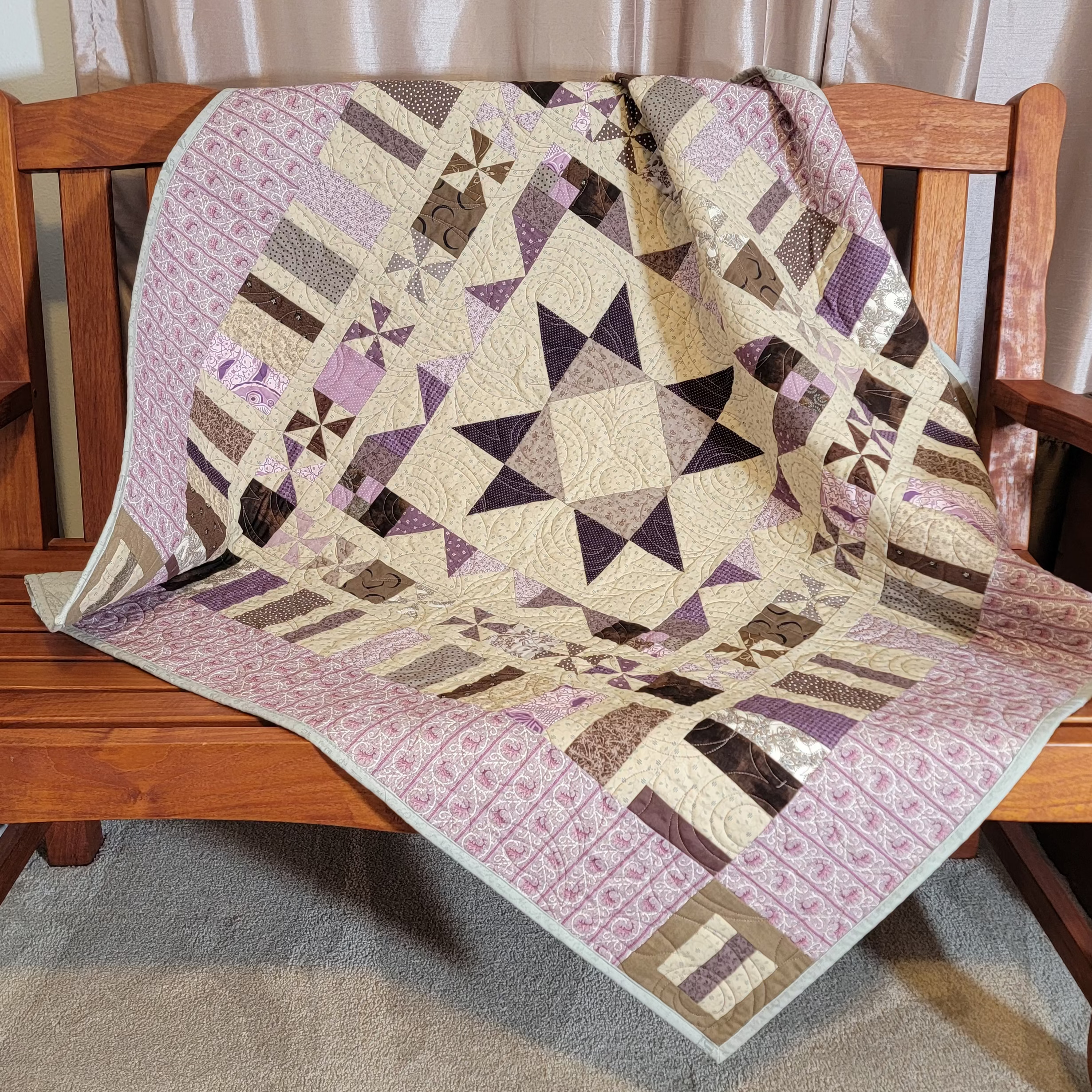 A geometric patchwork quilt in shades of lavender, beige, and brown draped over a wooden bench in a home setting.