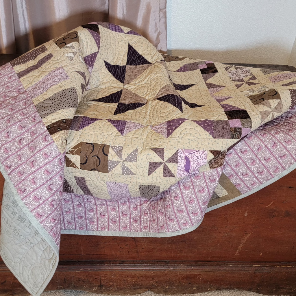 A handmade quilted patchwork blanket with geometric and windmill patterns in beige, purple, and brown, draped over a rustic wooden chest in an indoor setting.