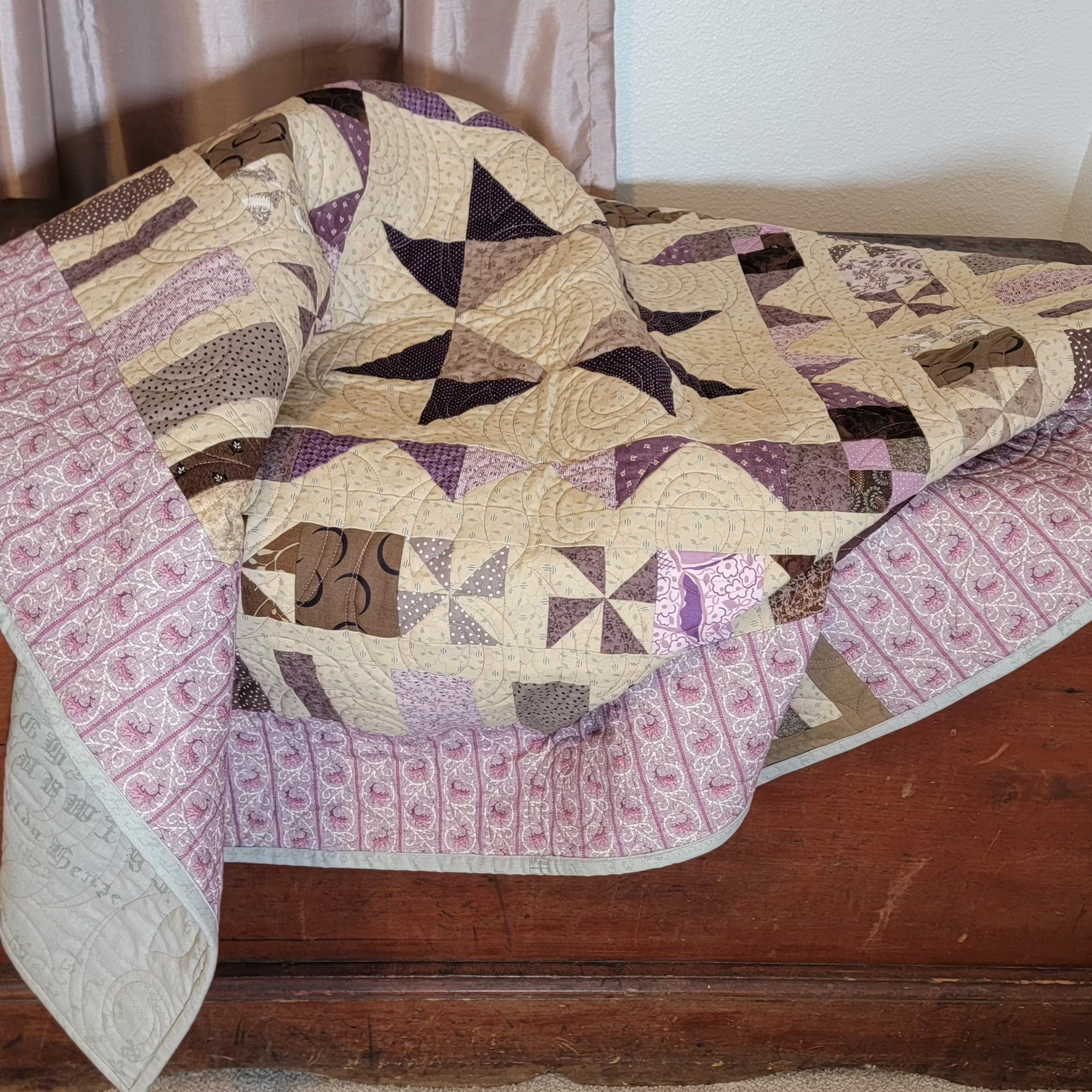 A handmade quilted patchwork blanket with geometric and windmill patterns in beige, purple, and brown, draped over a rustic wooden chest in an indoor setting.
