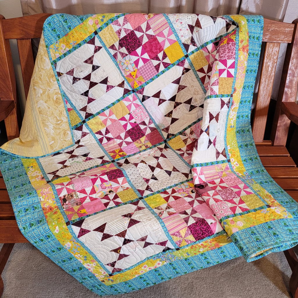 A colorful patchwork quilt draped over a wooden bench in a home setting, featuring geometric and floral block patterns in pink, yellow, teal, and white.