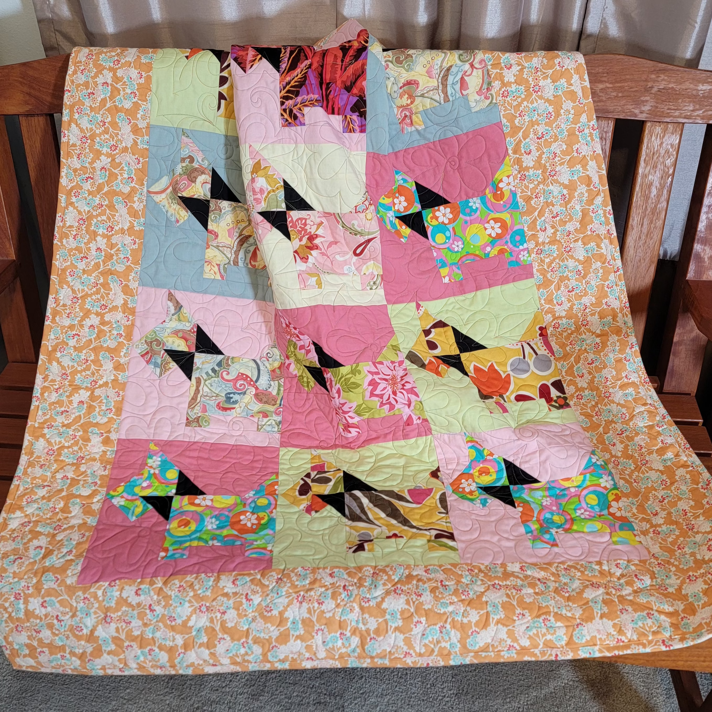 A colorful patchwork quilt draped over a wooden bench in a home setting. The quilt features a variety of floral and geometric patterns in the shape of scottie dogs in pink, orange, green, blue, and yellow, with a matching orange and white floral border.