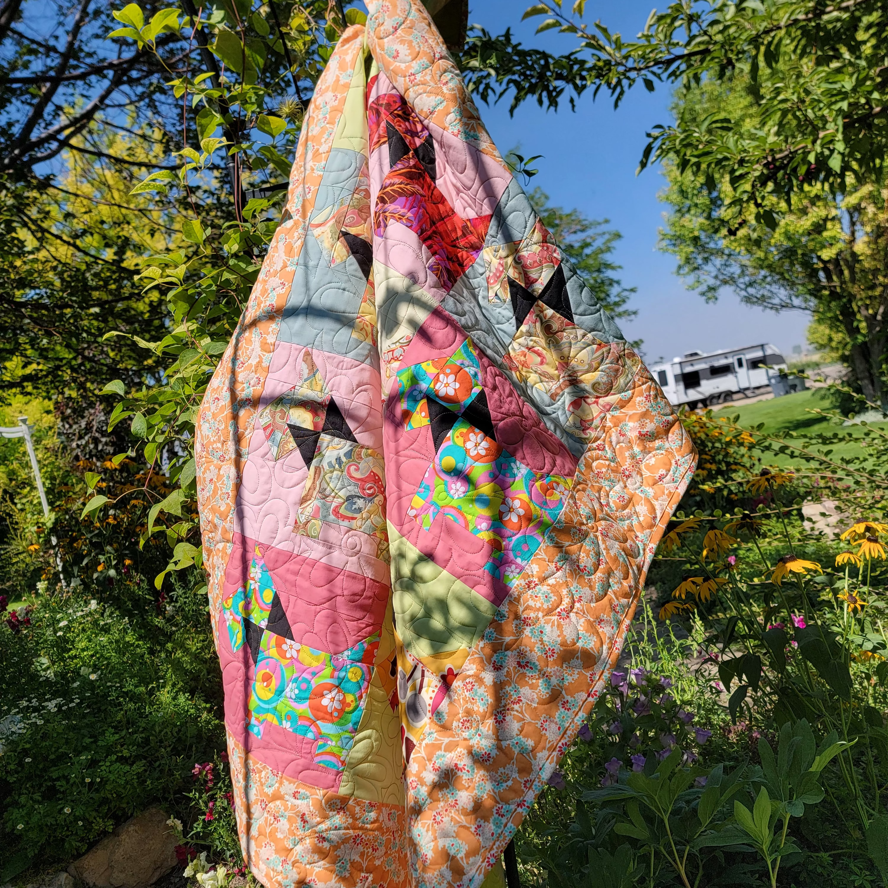 A colorful handmade quilt with a patchwork design, hanging outdoors from a wooden post in a garden with trees, flowers, and a blue sky in the background.