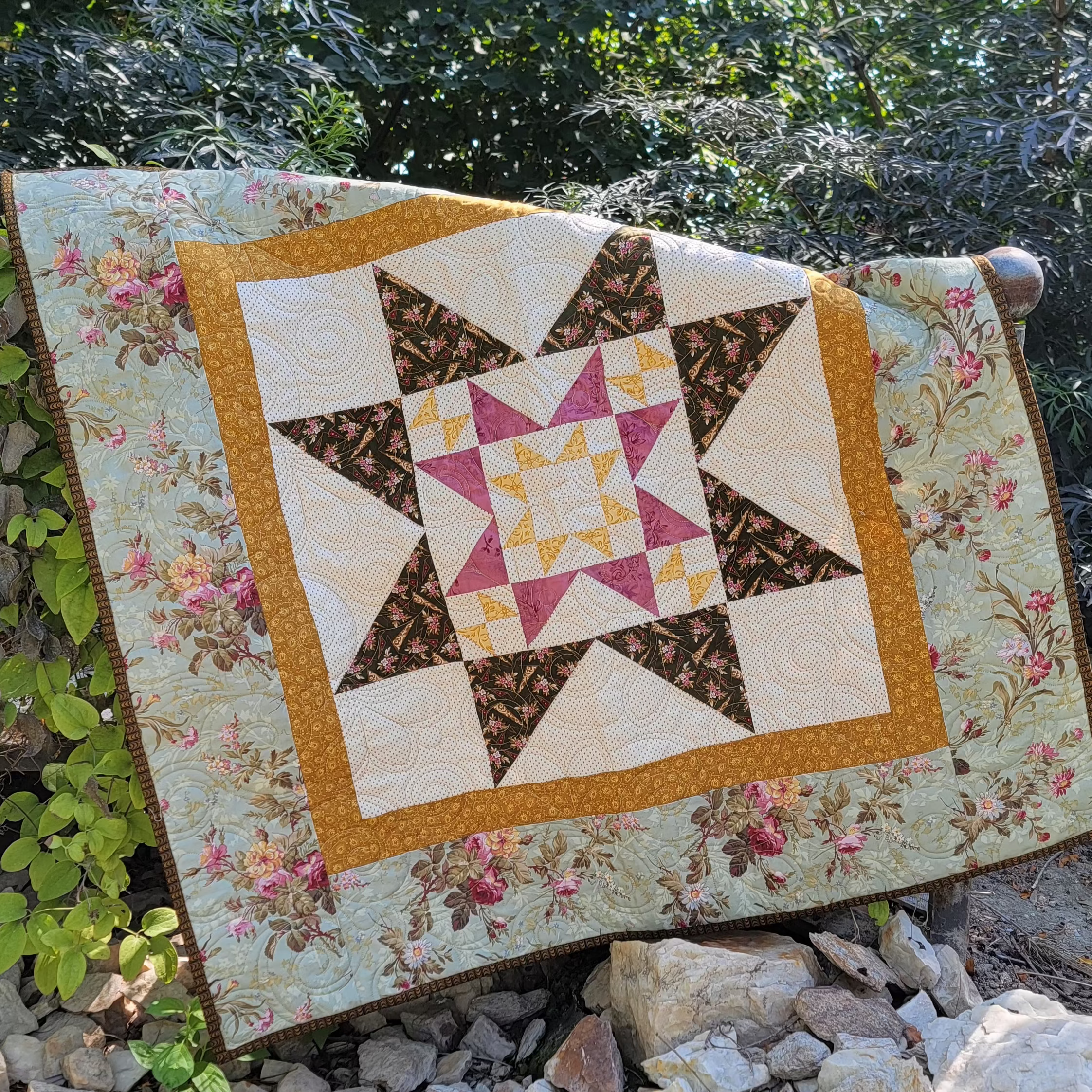 A handmade quilted blanket with a geometric star pattern and floral borders is draped outdoors over rocks with green foliage in the background.