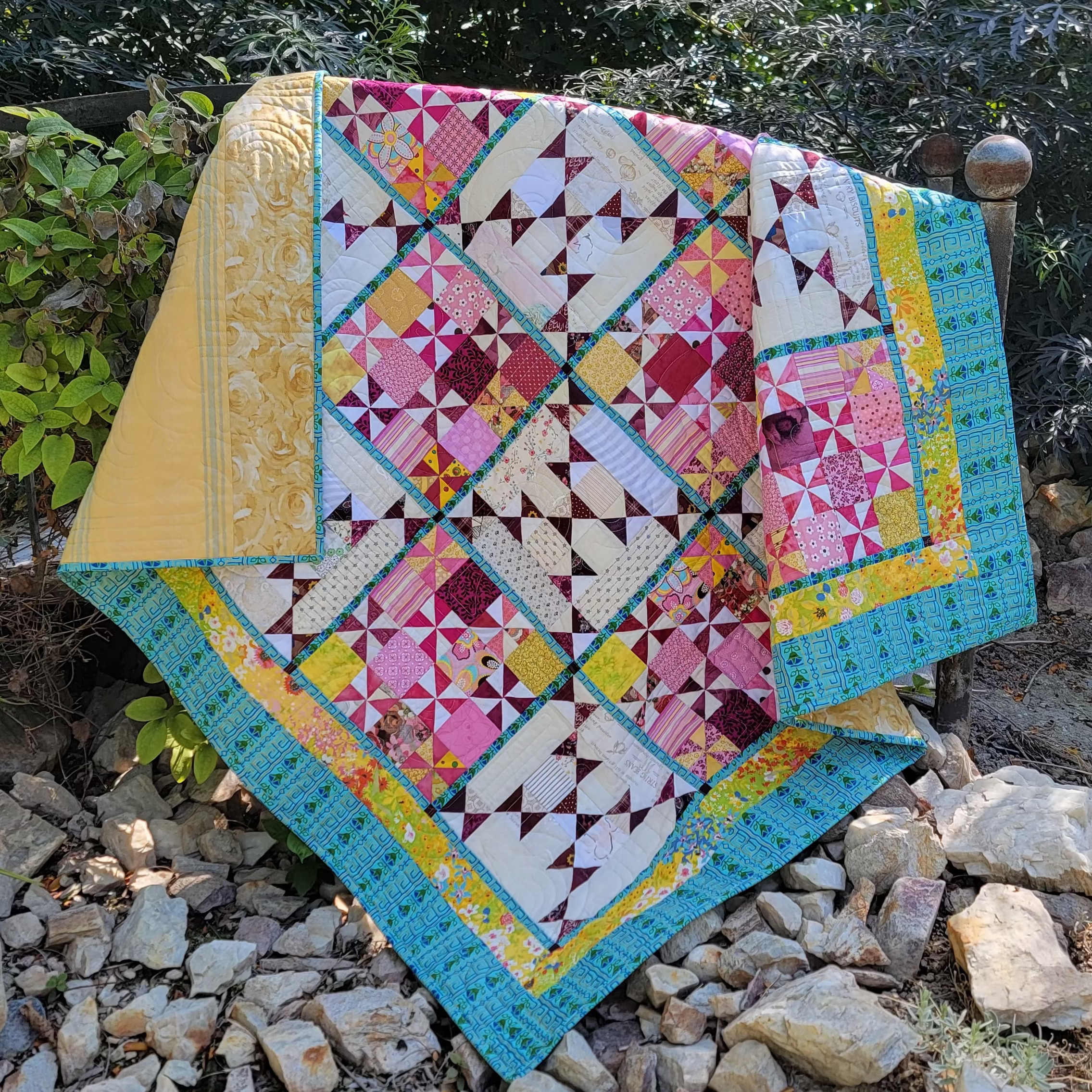 A colorful patchwork quilt draped over a wooden post outdoors on a rocky path with green foliage in the background. The quilt features a geometric diamond pattern with pink, teal, yellow, and white sections.
