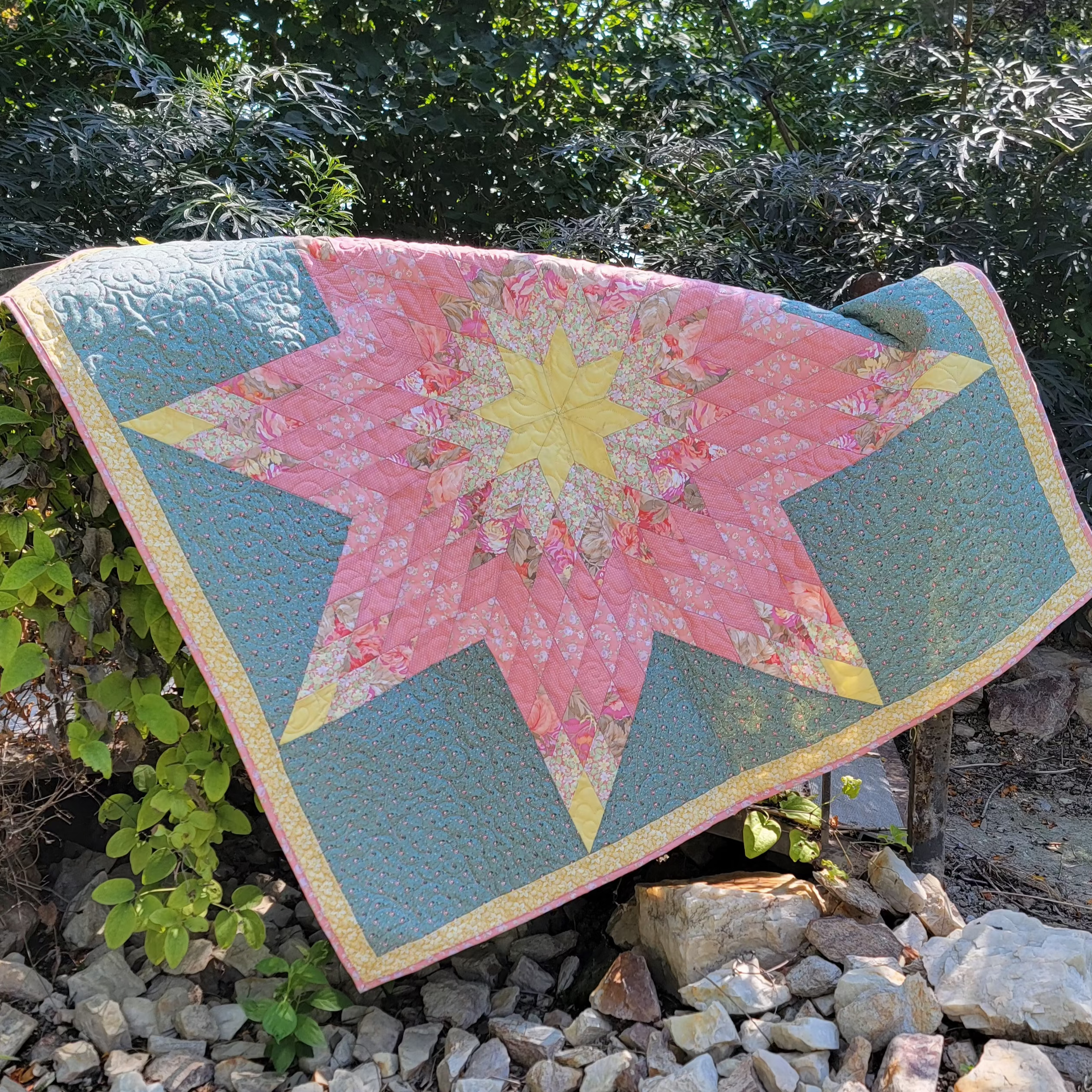 A handmade quilted starburst quilt with pink floral sections, teal blue corner panels, and a yellow center, draped outdoors over rocks with green foliage in the background.