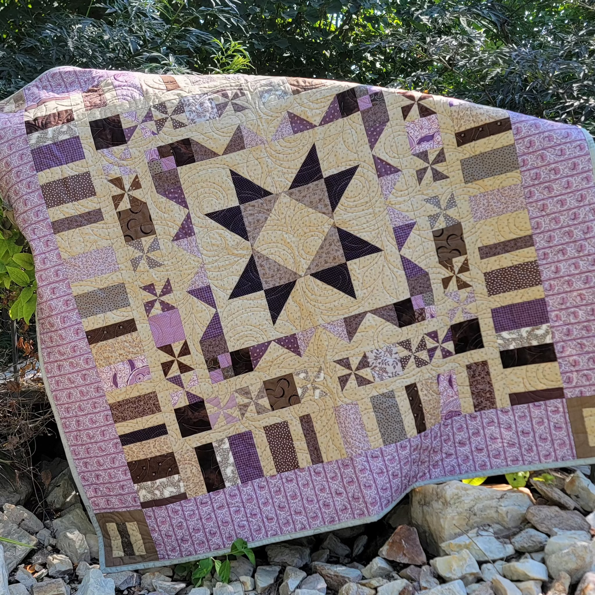 A handmade patchwork quilt with a geometric star and block pattern in shades of purple, beige, and brown, draped over a rustic wooden bridge in an outdoor garden setting with rocks and greenery.