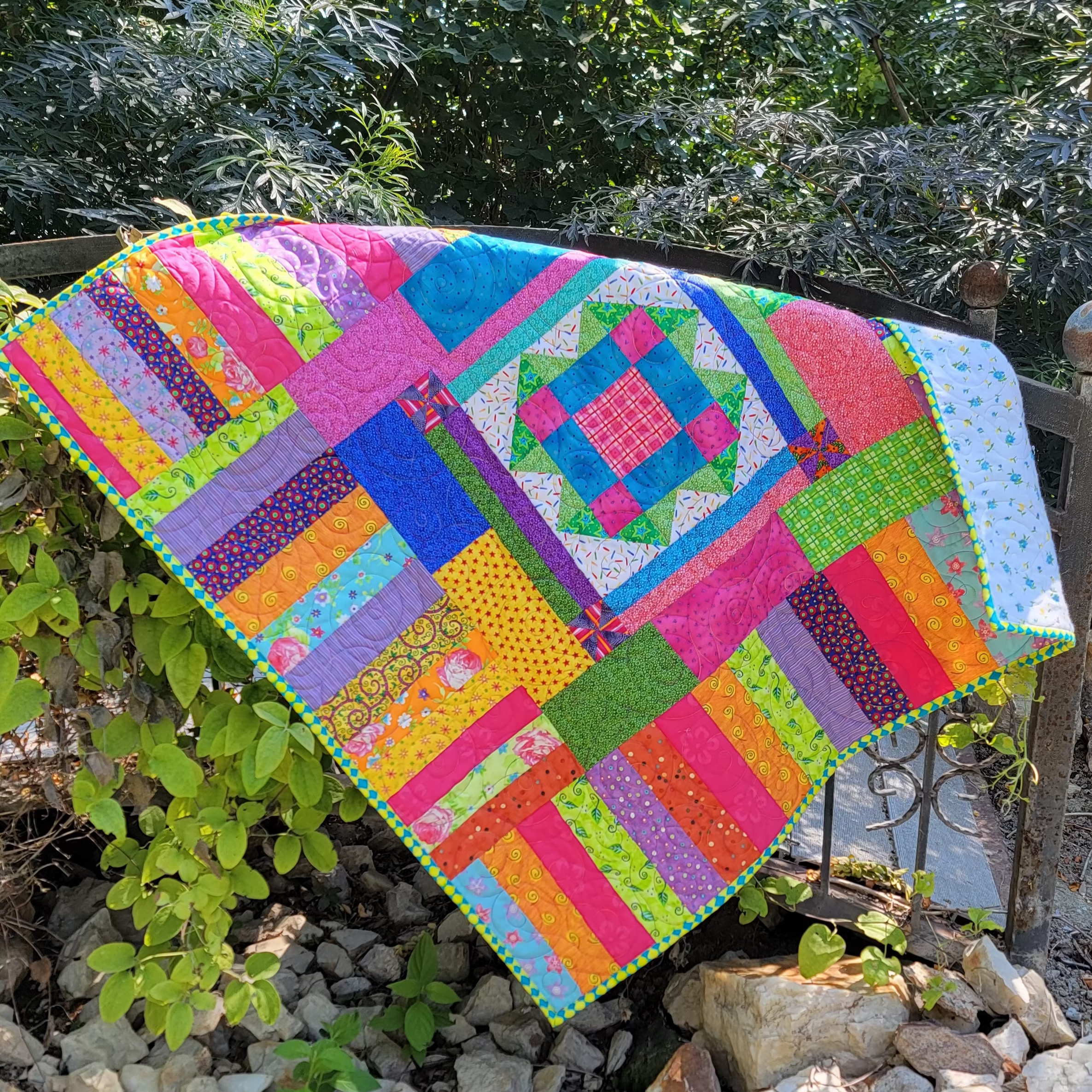 A colorful patchwork quilted blanket with a geometric diamond pattern is draped over a wooden bridge in an outdoor setting.