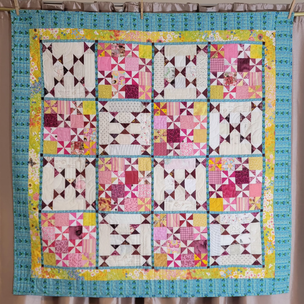 A handmade patchwork quilt hanging on a rod with wooden clothespins against a beige curtain backdrop. The quilt features a colorful grid of pink, purple, yellow, and white geometric blocks, bordered by a turquoise blue patterned section and a yellow floral inner border.