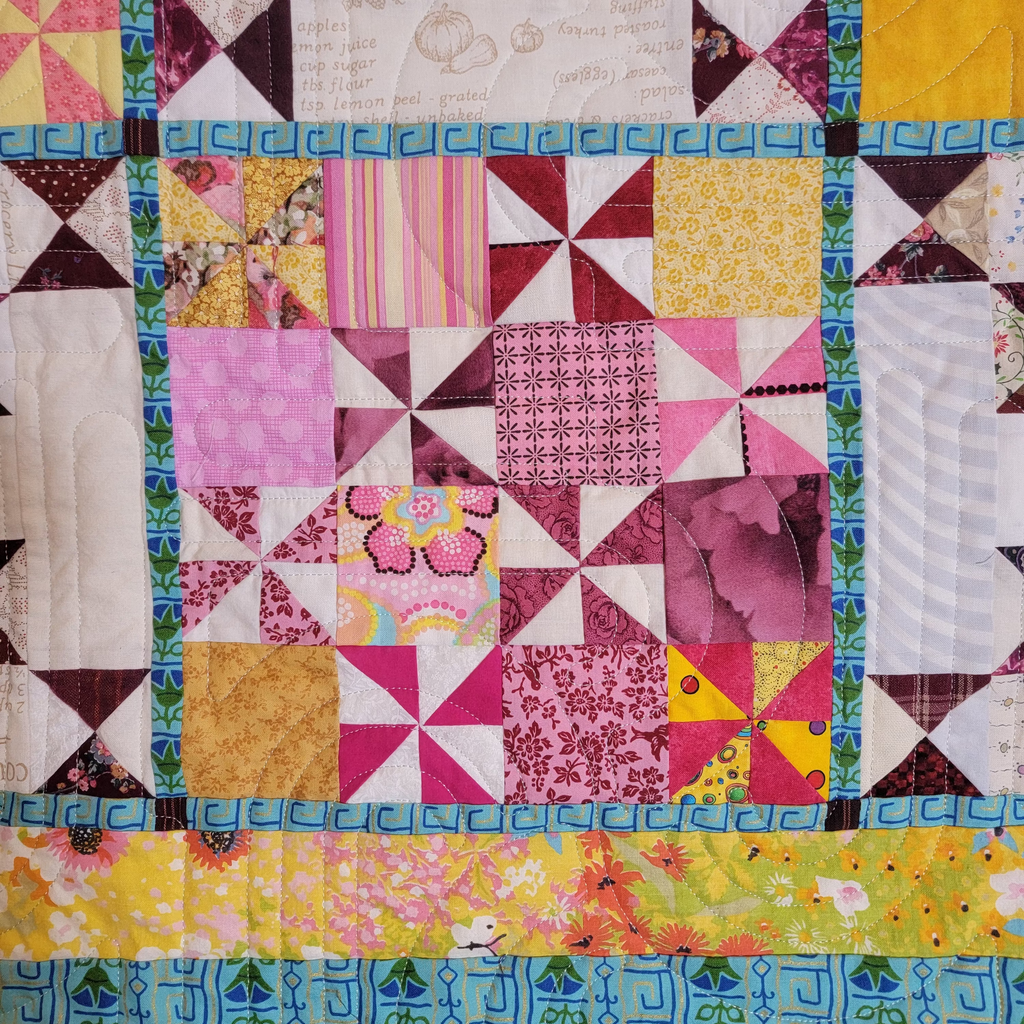 A close-up of a colorful patchwork quilt featuring various fabric blocks with geometric and floral patterns in pink, yellow, blue, green, and white.