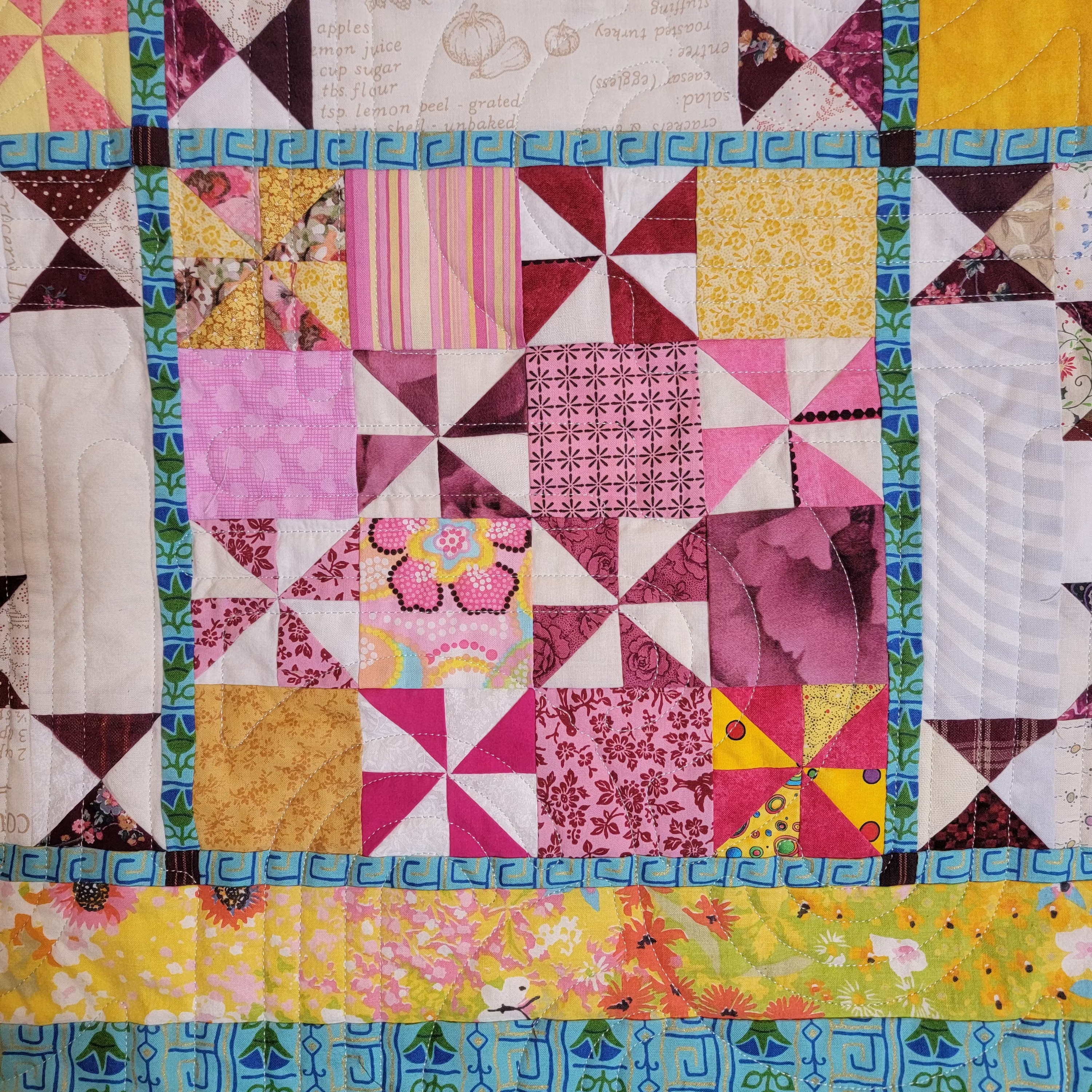 A close-up of a colorful patchwork quilt featuring various fabric blocks with geometric and floral patterns in pink, yellow, blue, green, and white.