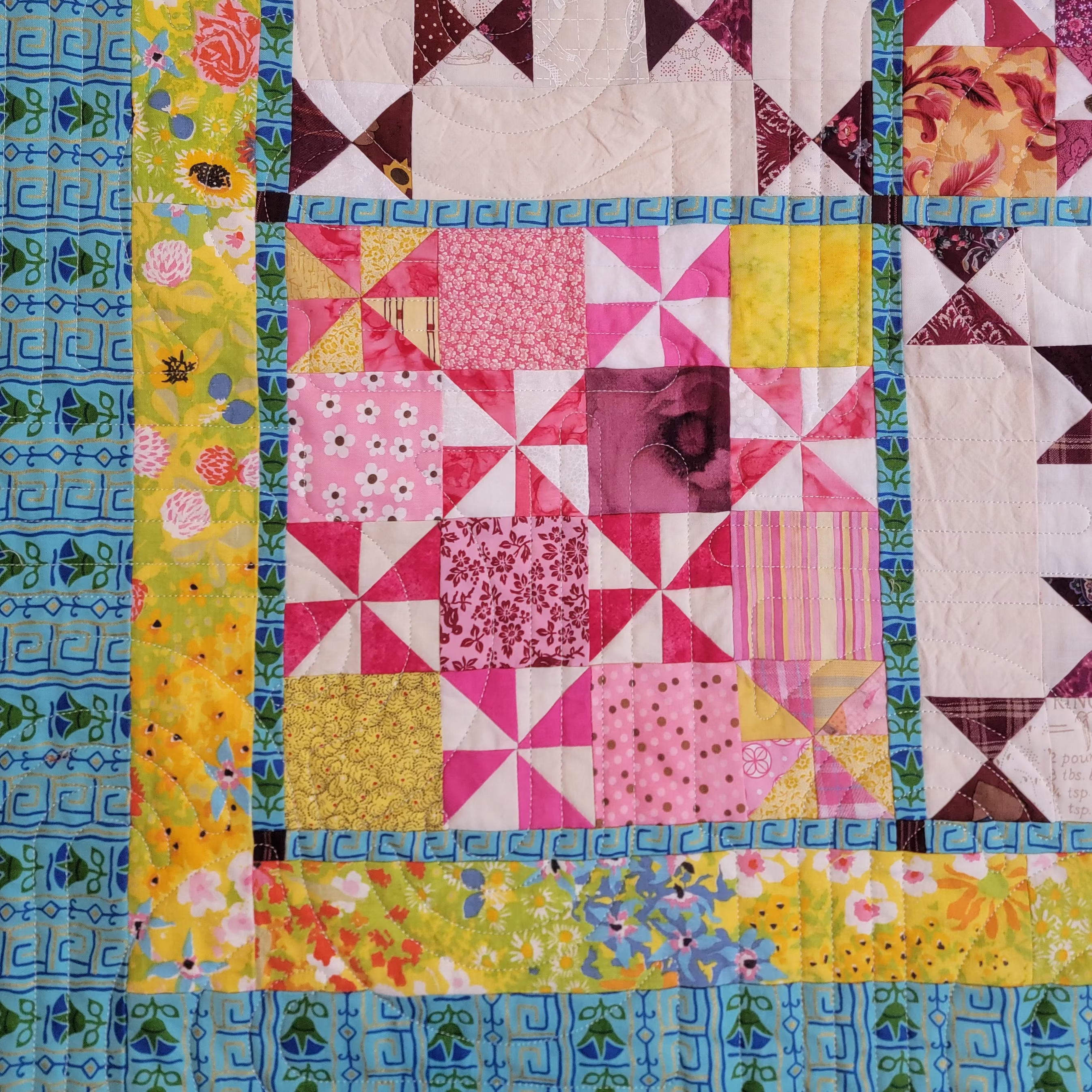 A close-up of a colorful quilted fabric panel with patchwork blocks in pink, yellow, white, and dark plum, bordered by patterned blue and yellow fabric strips.