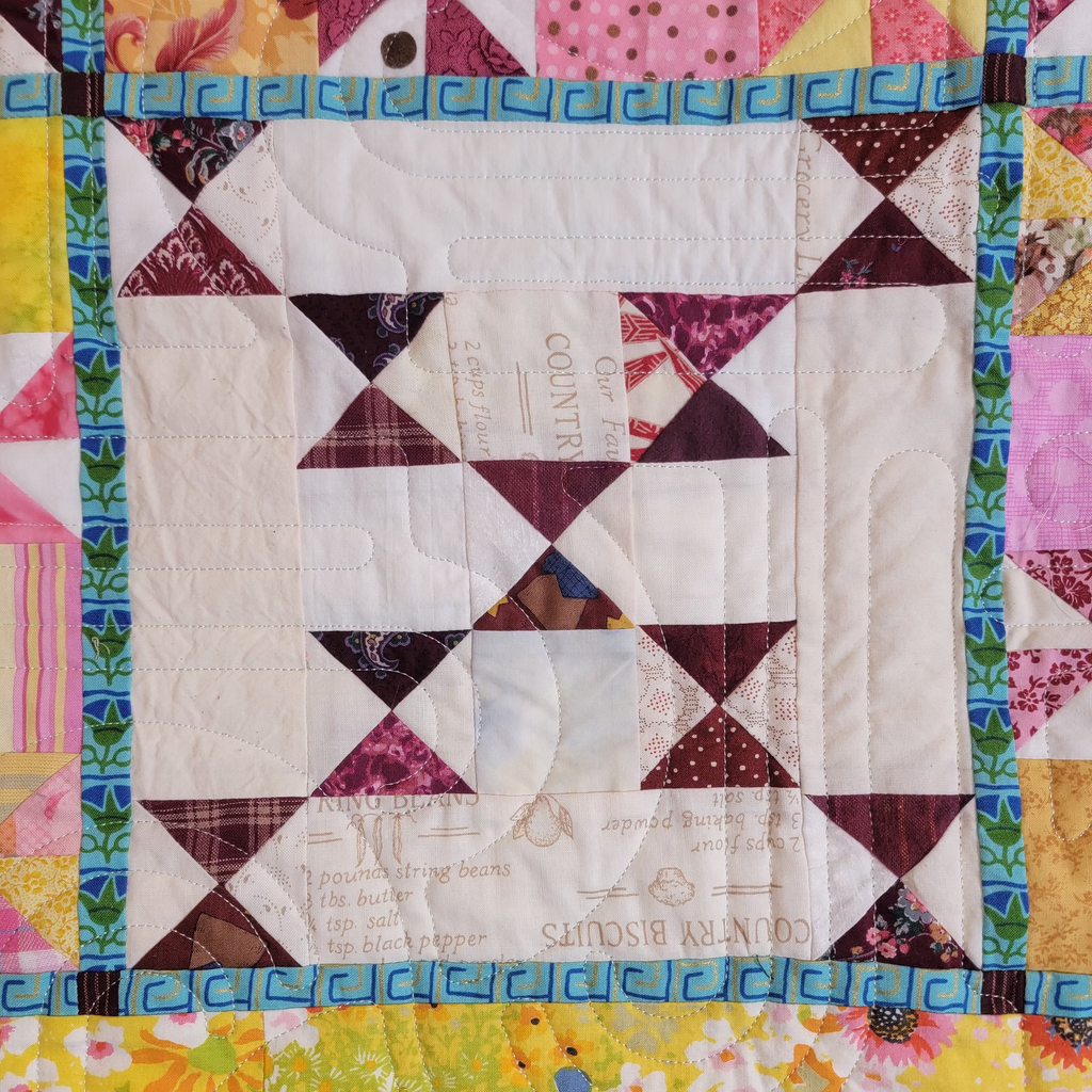 A close-up of a colorful quilted fabric featuring a central geometric pattern made of dark red, white, and brown triangles, surrounded by a vibrant multicolor border with floral and geometric prints.
