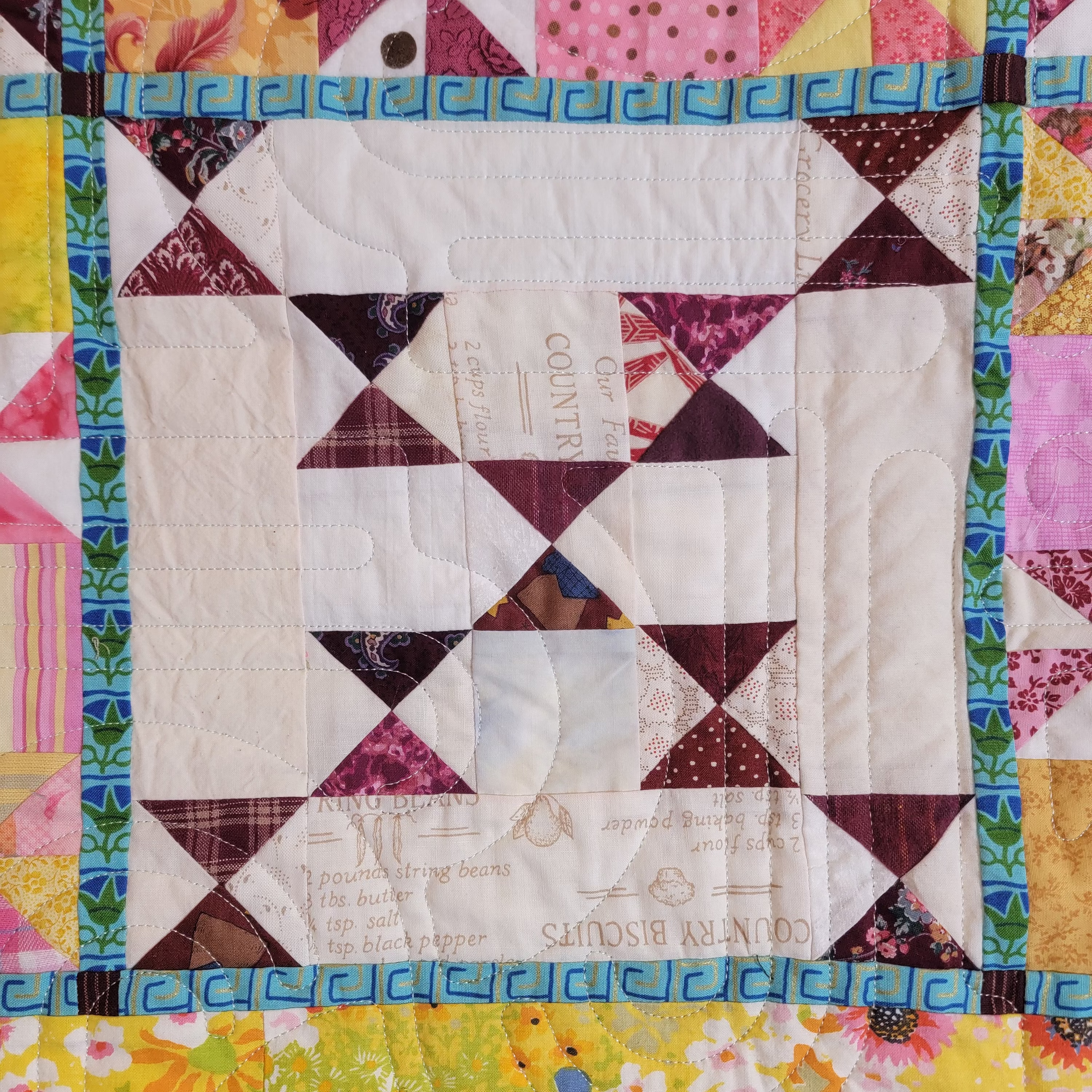 A close-up of a colorful quilted fabric featuring a central geometric pattern made of dark red, white, and brown triangles, surrounded by a vibrant multicolor border with floral and geometric prints.
