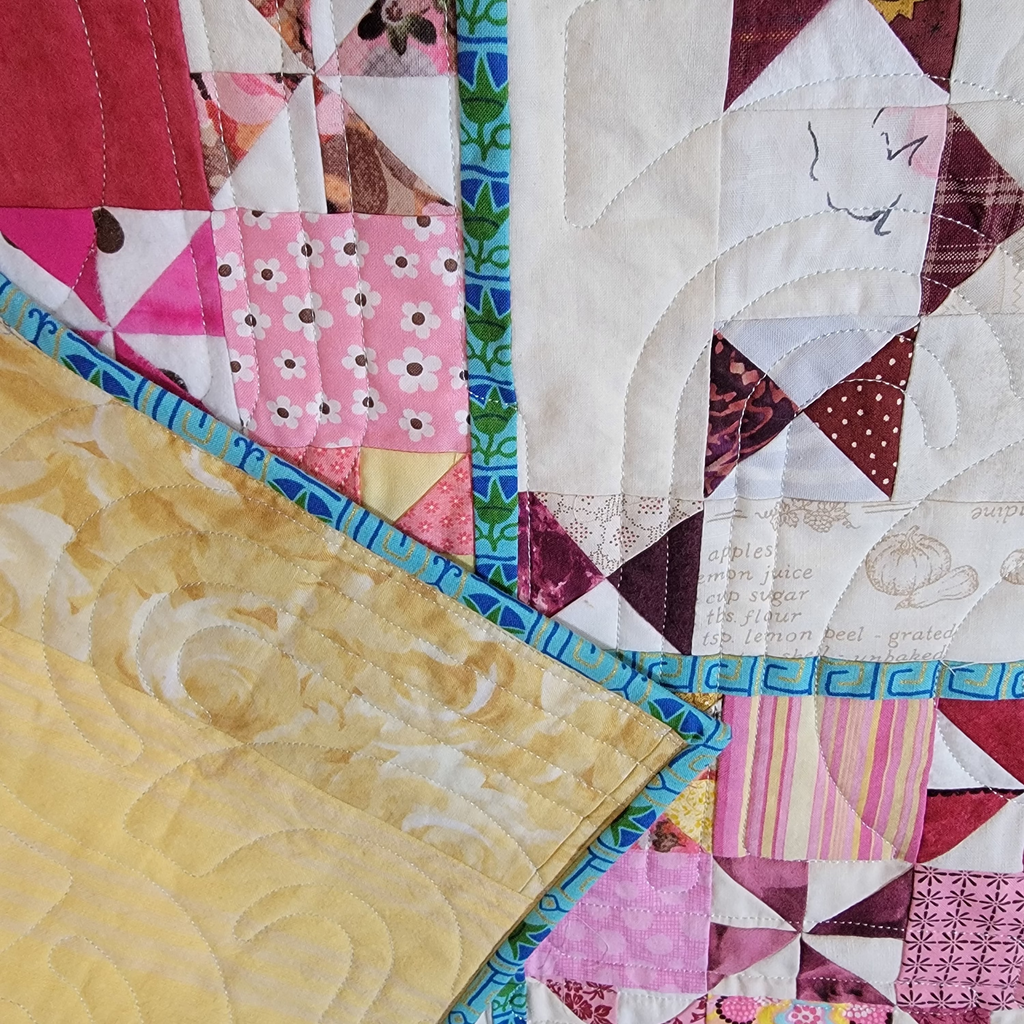 A close-up of a colorful patchwork quilt with various geometric and floral fabric blocks, featuring pink, red, yellow, and white patterns with blue borders.