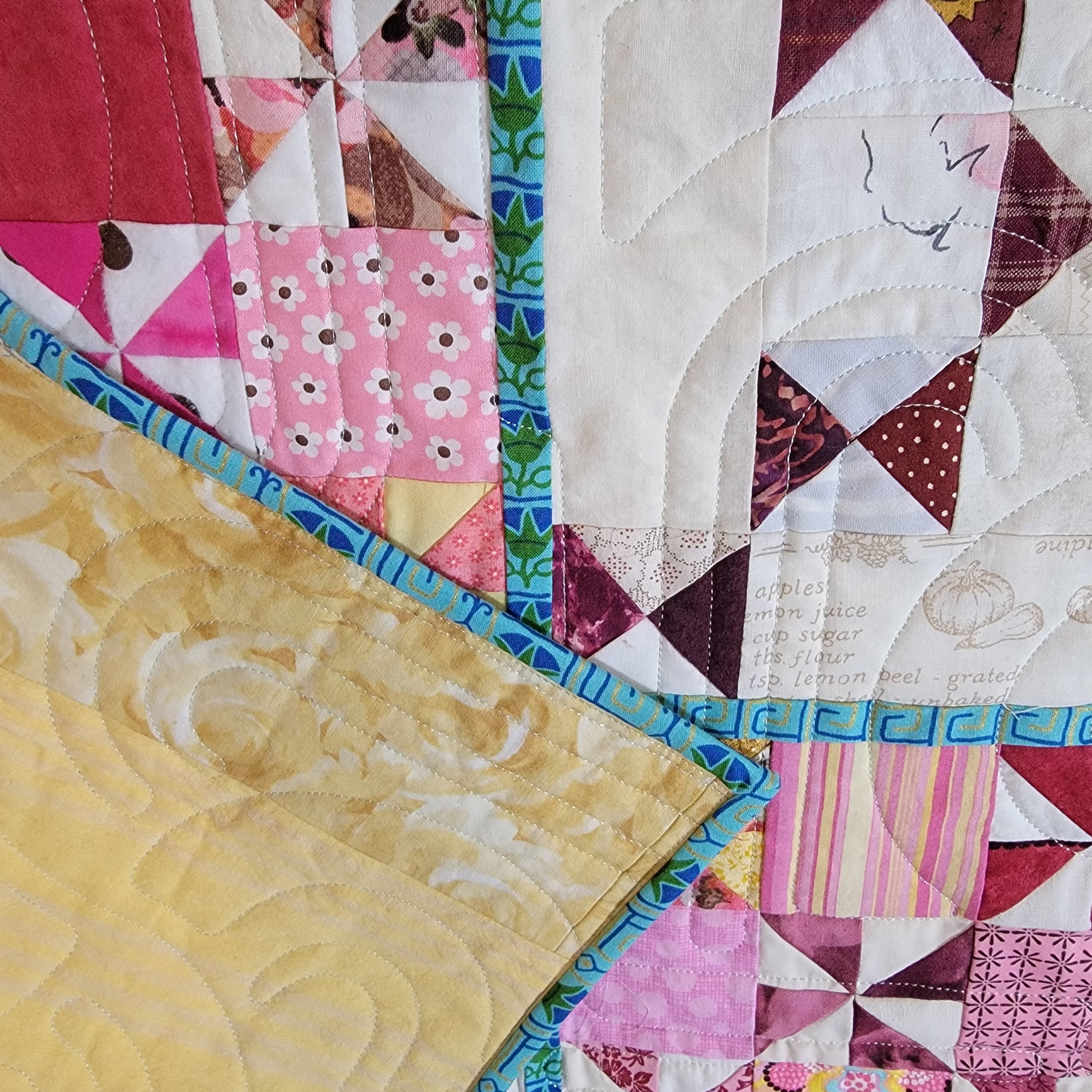 A close-up of a colorful patchwork quilt with various geometric and floral fabric blocks, featuring pink, red, yellow, and white patterns with blue borders.