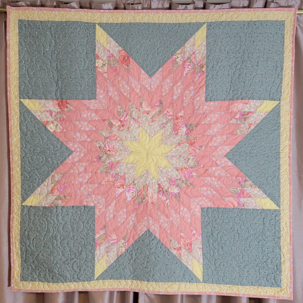A handmade quilted starburst quilt hanging on a rod against a curtain backdrop. The quilt features a central pink floral star with a yellow center, surrounded by yellow triangles and four muted sage green corners.